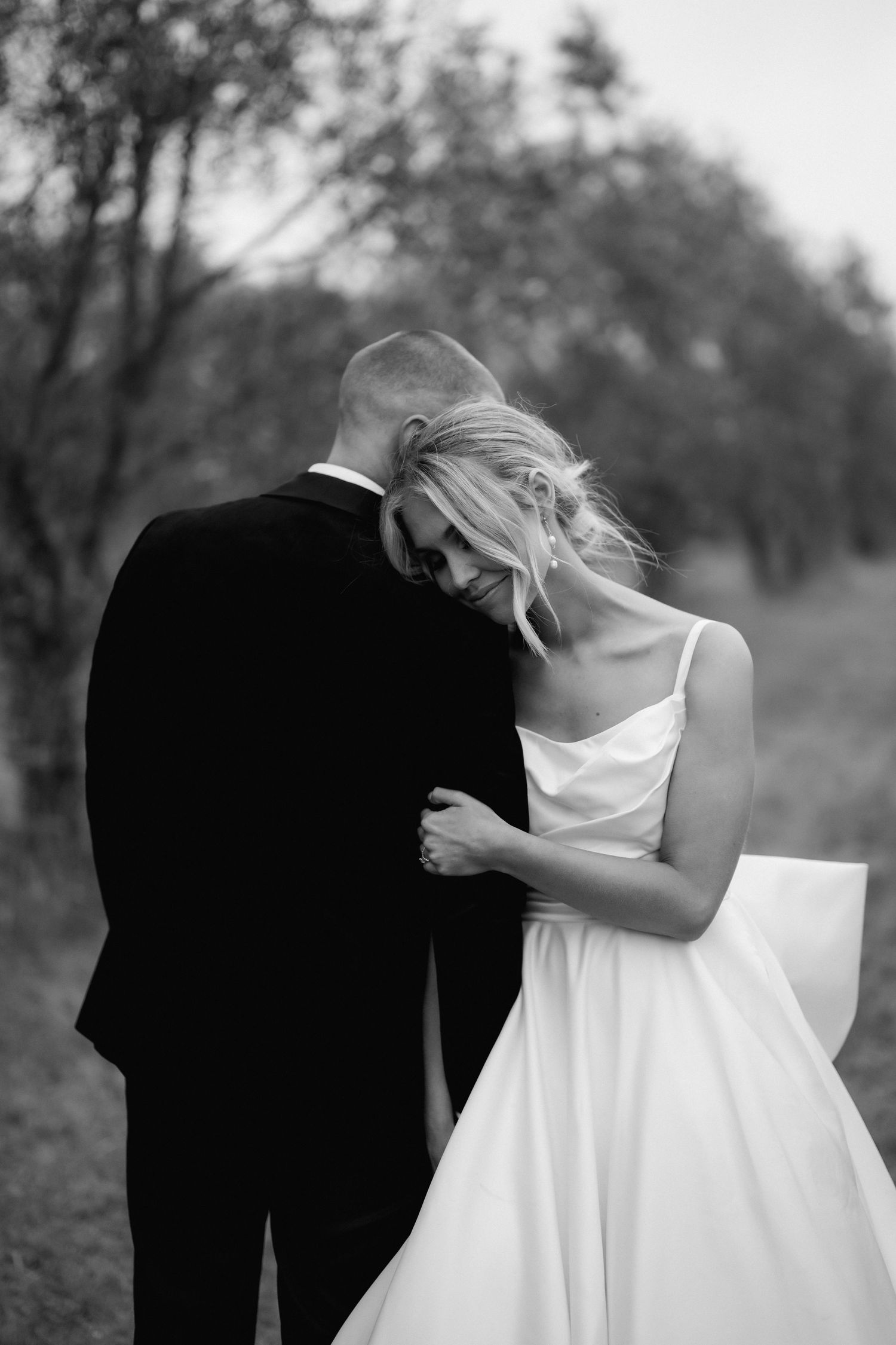 Black and white photo of a bride in a white gown leaning on a groom in a black suit outdoors.