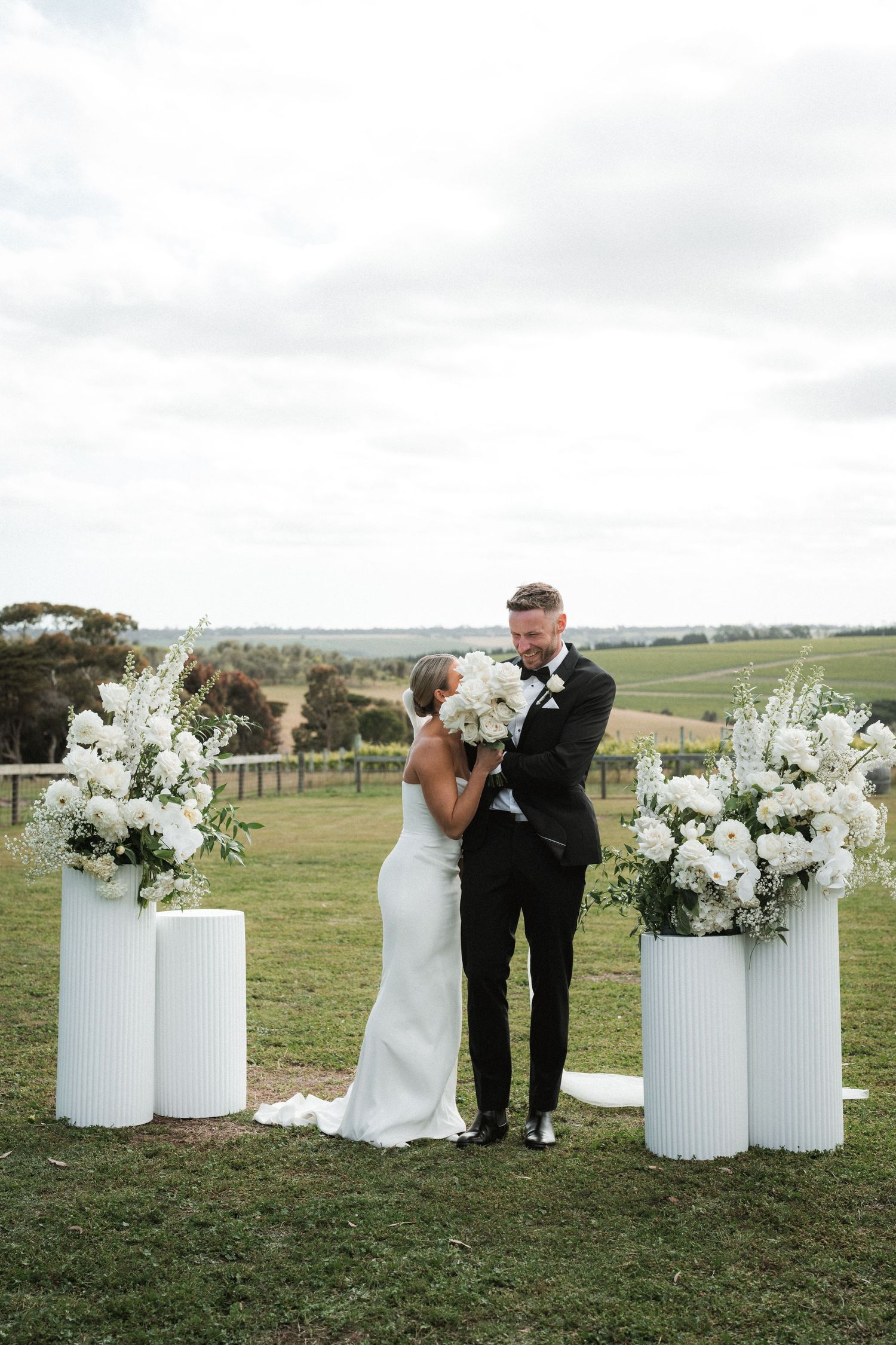 Couple embracing between tall white floral arrangements during outdoor ceremony.