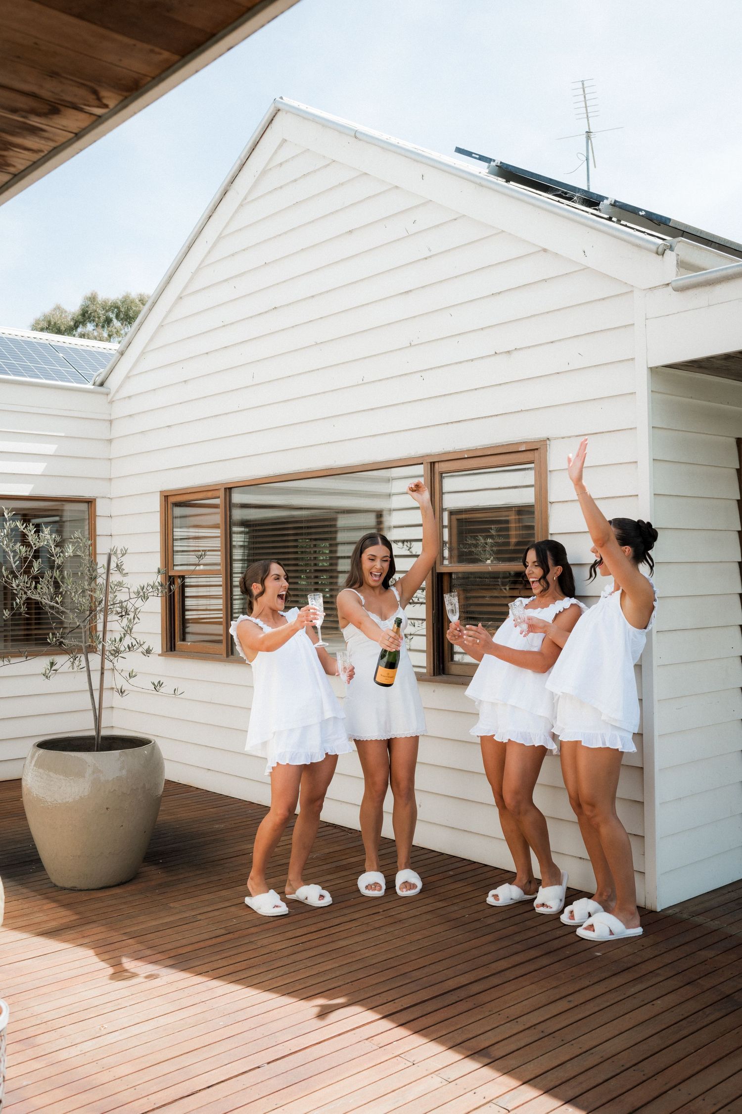 Group wearing white robes and slippers pose playfully on a wooden deck of a modern spa.
