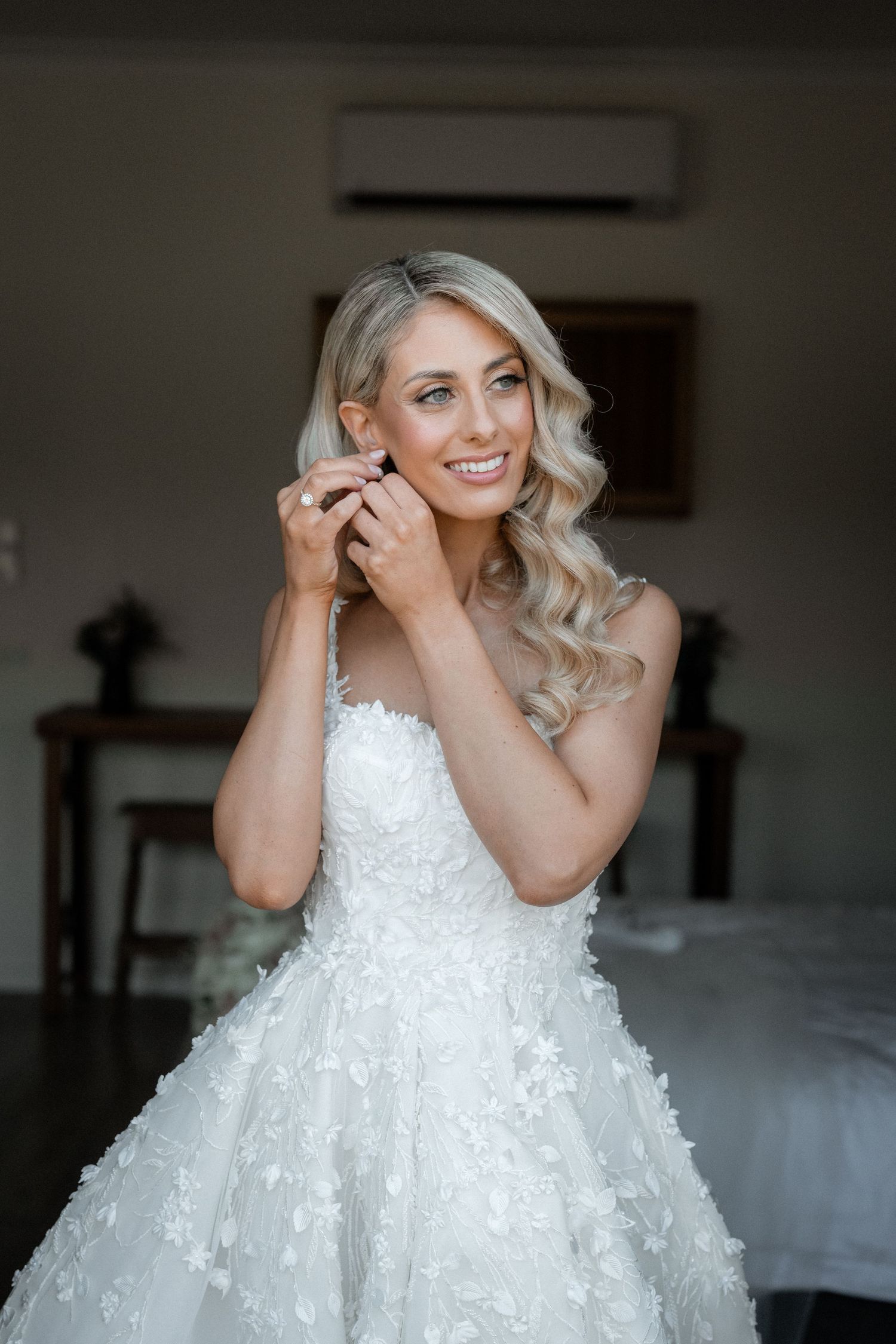 A beautiful bride getting ready in a stunning white lacy ball gown while putting on earrings.
