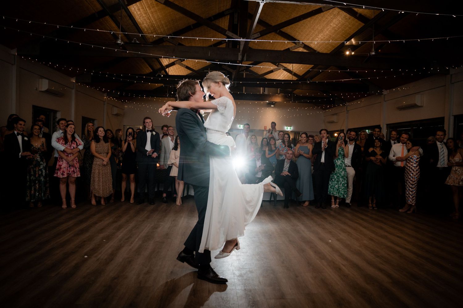 A couple shares their first dance under spotlights while guests watch from the edges of a rustic reception hall.