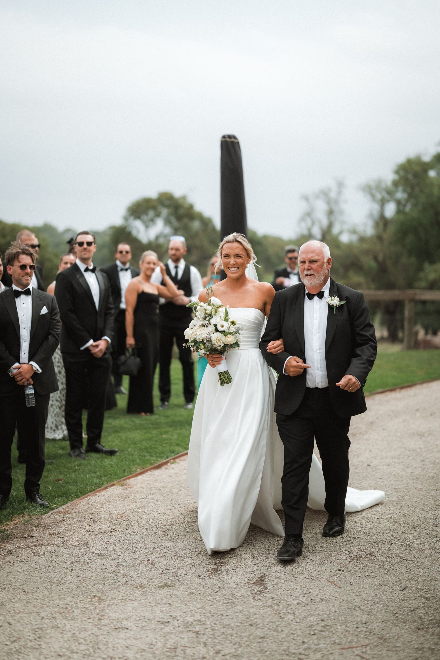 Wedding ceremony procession on a garden pathway with formal attire and floral bouquet.