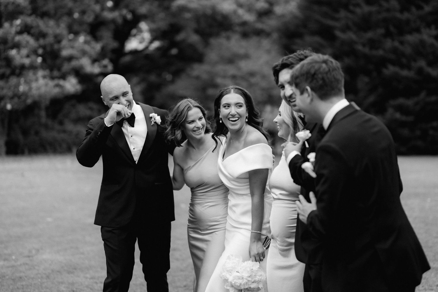 Wedding party members sharing candid laughter together outdoors on a grassy lawn.