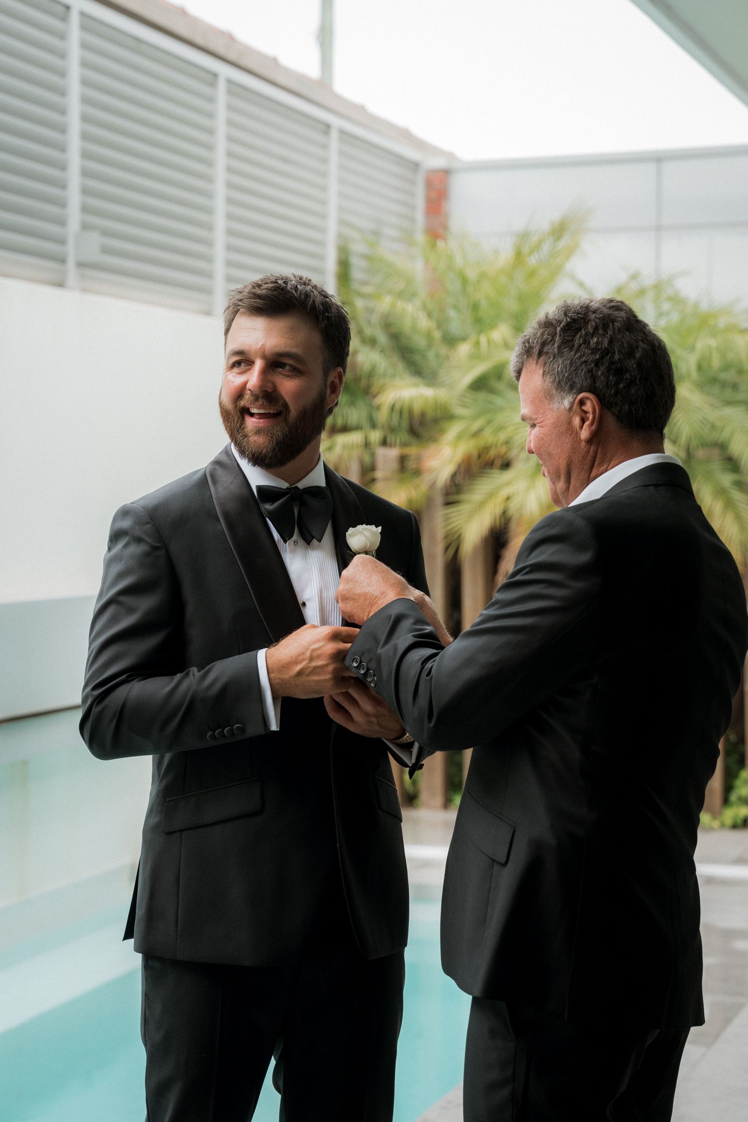 Two grooms in black tuxedos share a joyful moment adjusting cufflinks poolside at their wedding.