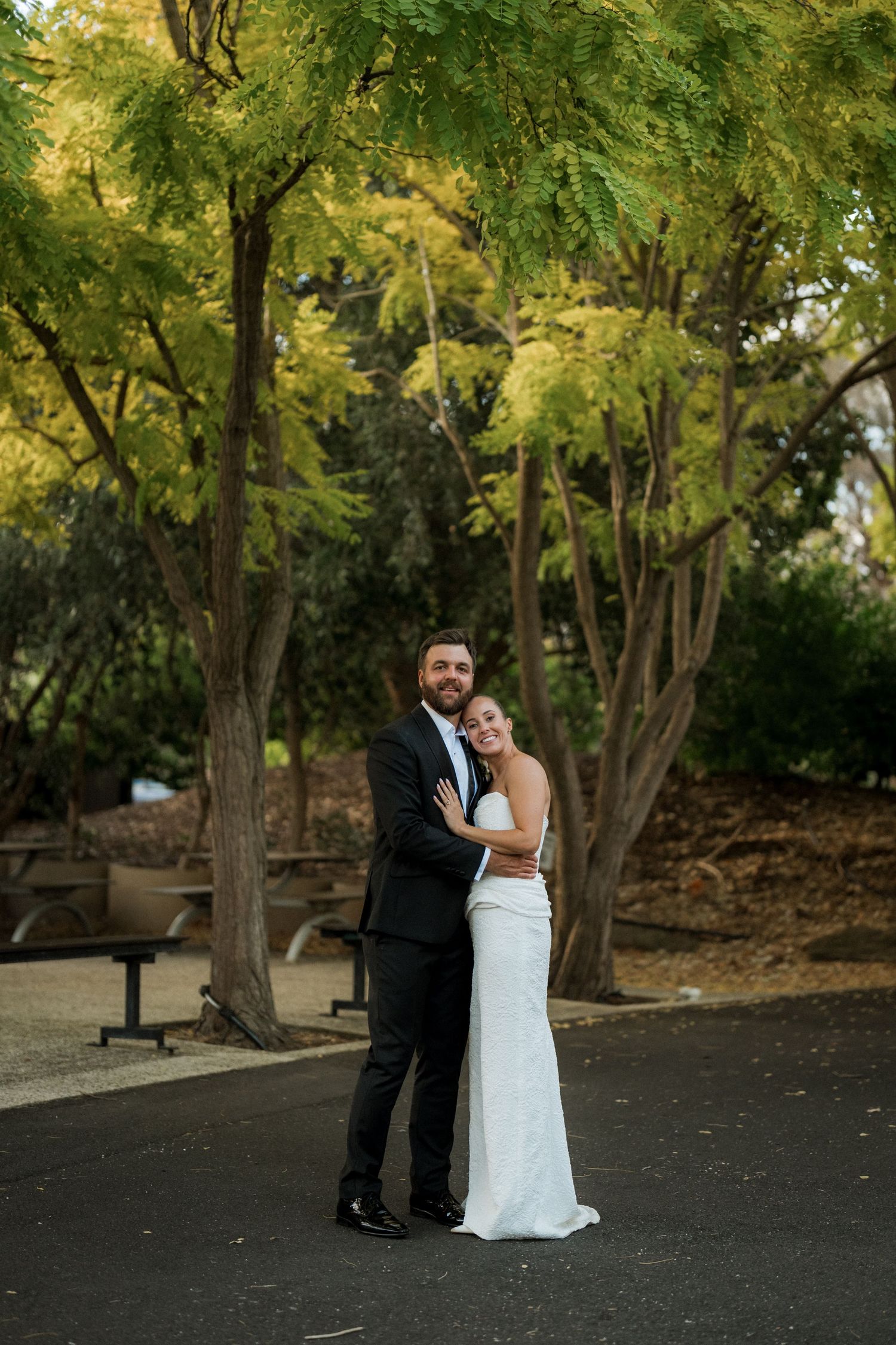 Bride and groom embracing outdoors surrounded by lush green trees on their wedding day.
