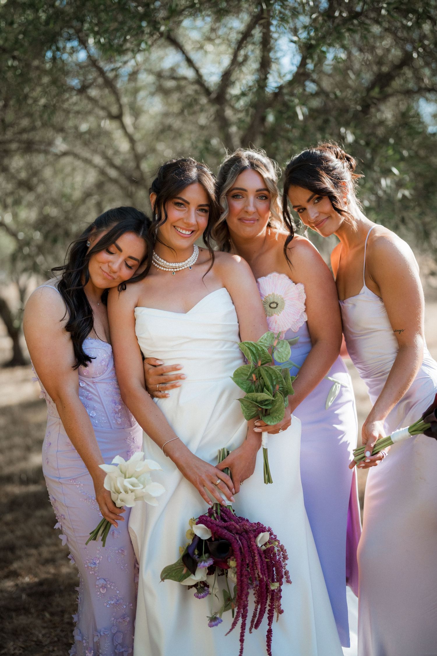 Bridesmaids in lavender dresses embrace bride during outdoor wedding photo session.