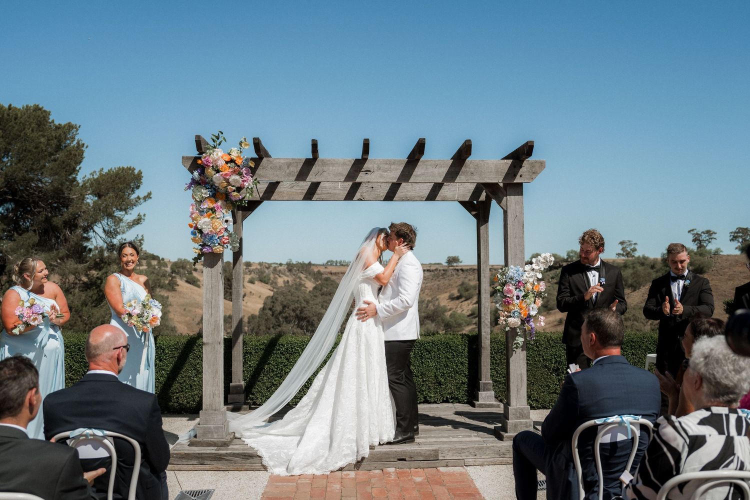 Bride and groom share first kiss under wooden pergola with floral arch at outdoor wedding ceremony.