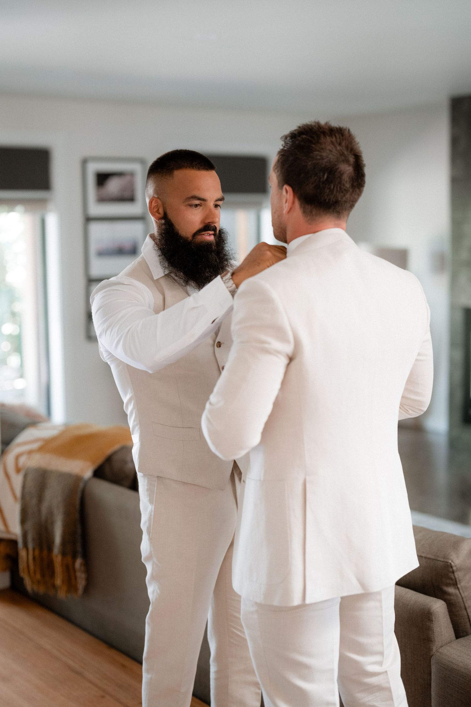 Two people in white formal attire share an intimate moment while getting ready indoors.