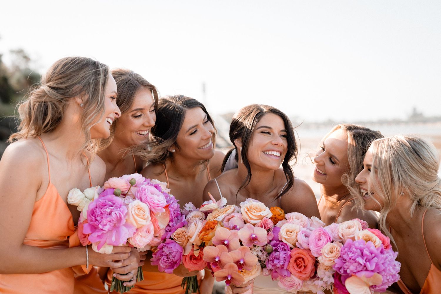 A row of bridesmaids holding vibrant pink and orange bouquets lean in close together for a group photo.