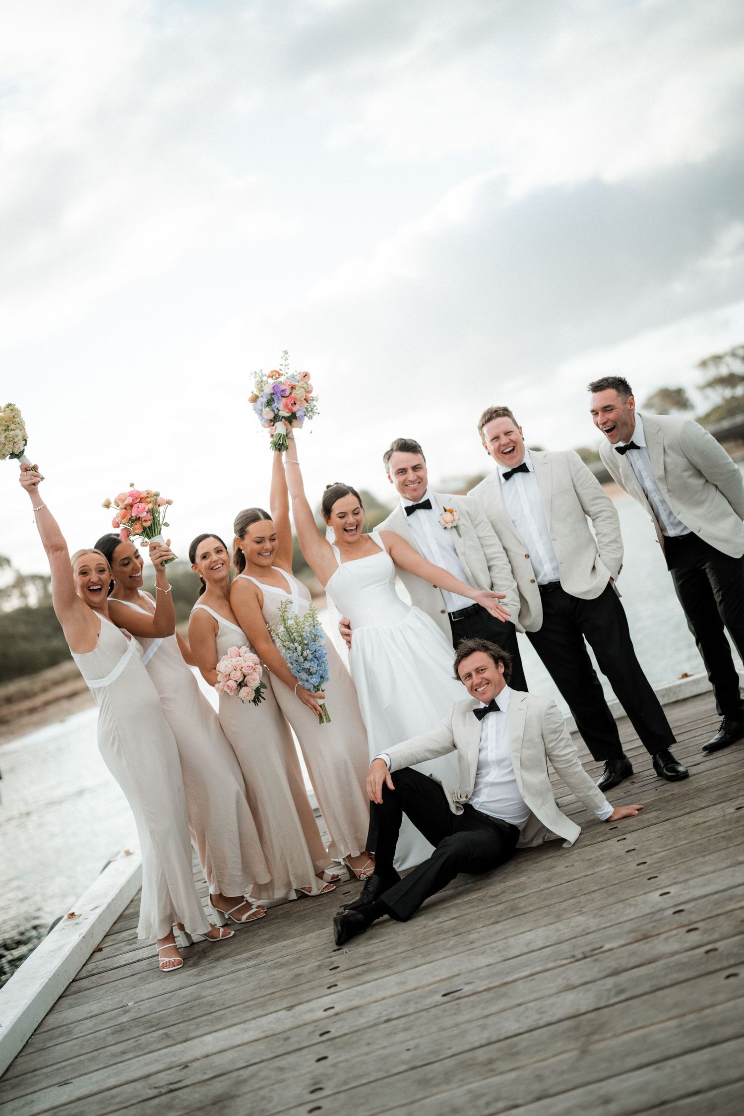 Wedding party poses playfully on a wooden dock by the water.