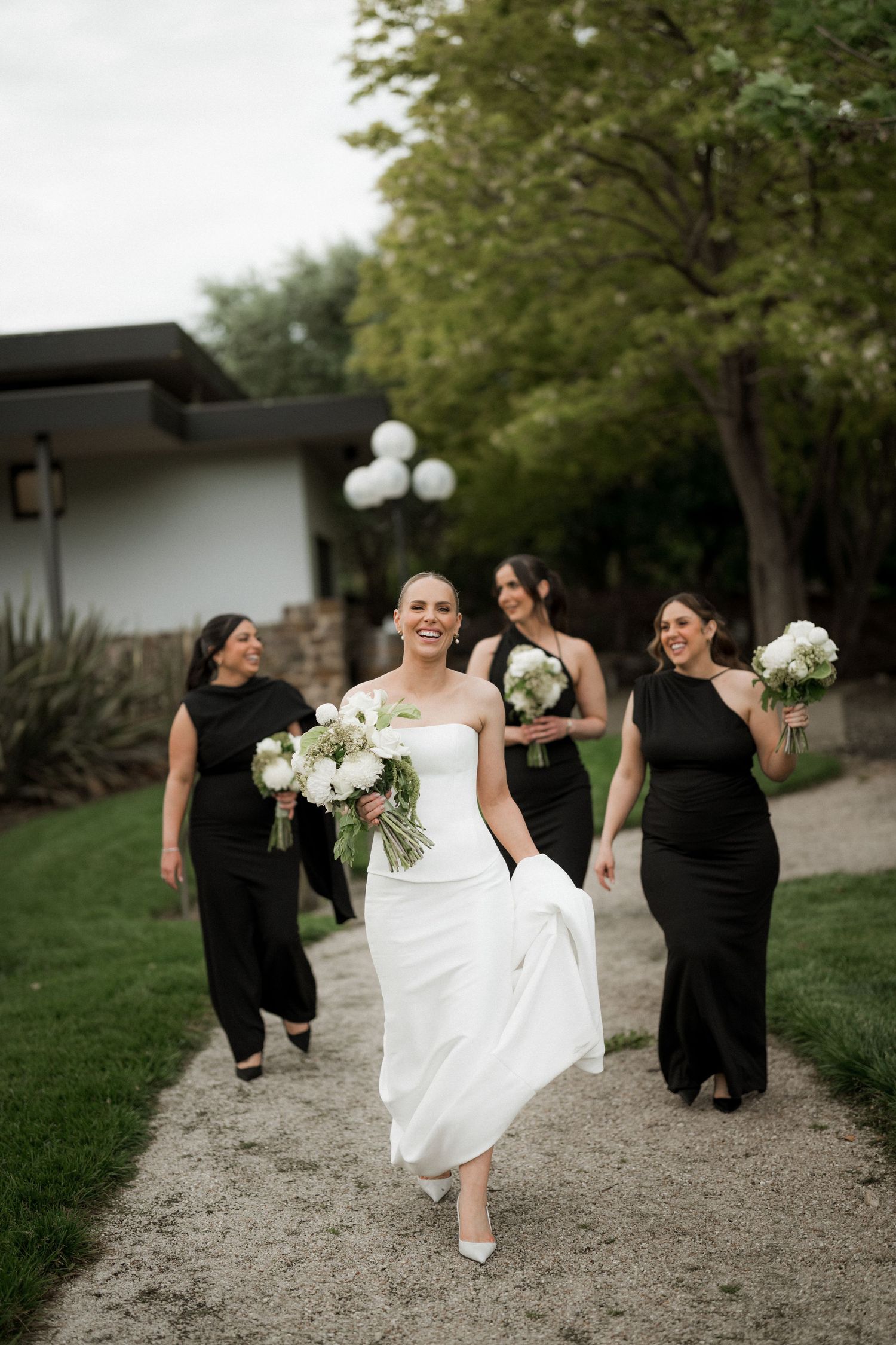 Wedding party walking and laughing along a garden path in black and white formal attire.