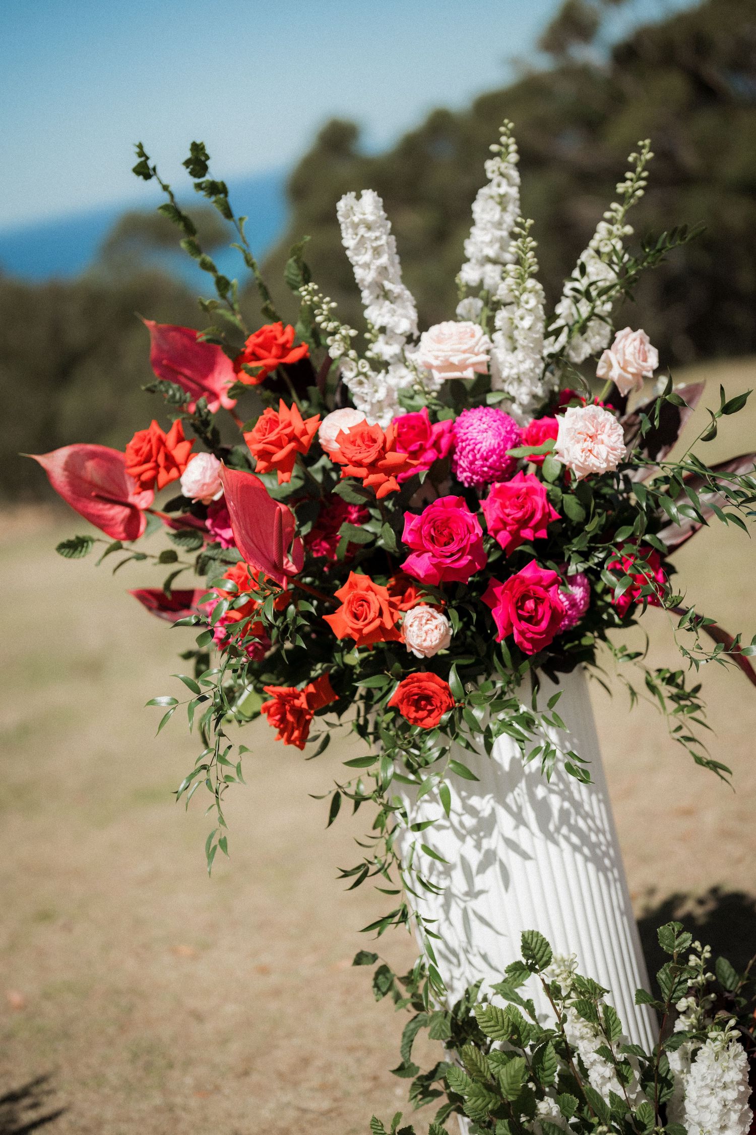 White pedestal vase with vibrant red, pink, and orange roses, anthurium, and white blooms outdoors at a wedding ceremony.