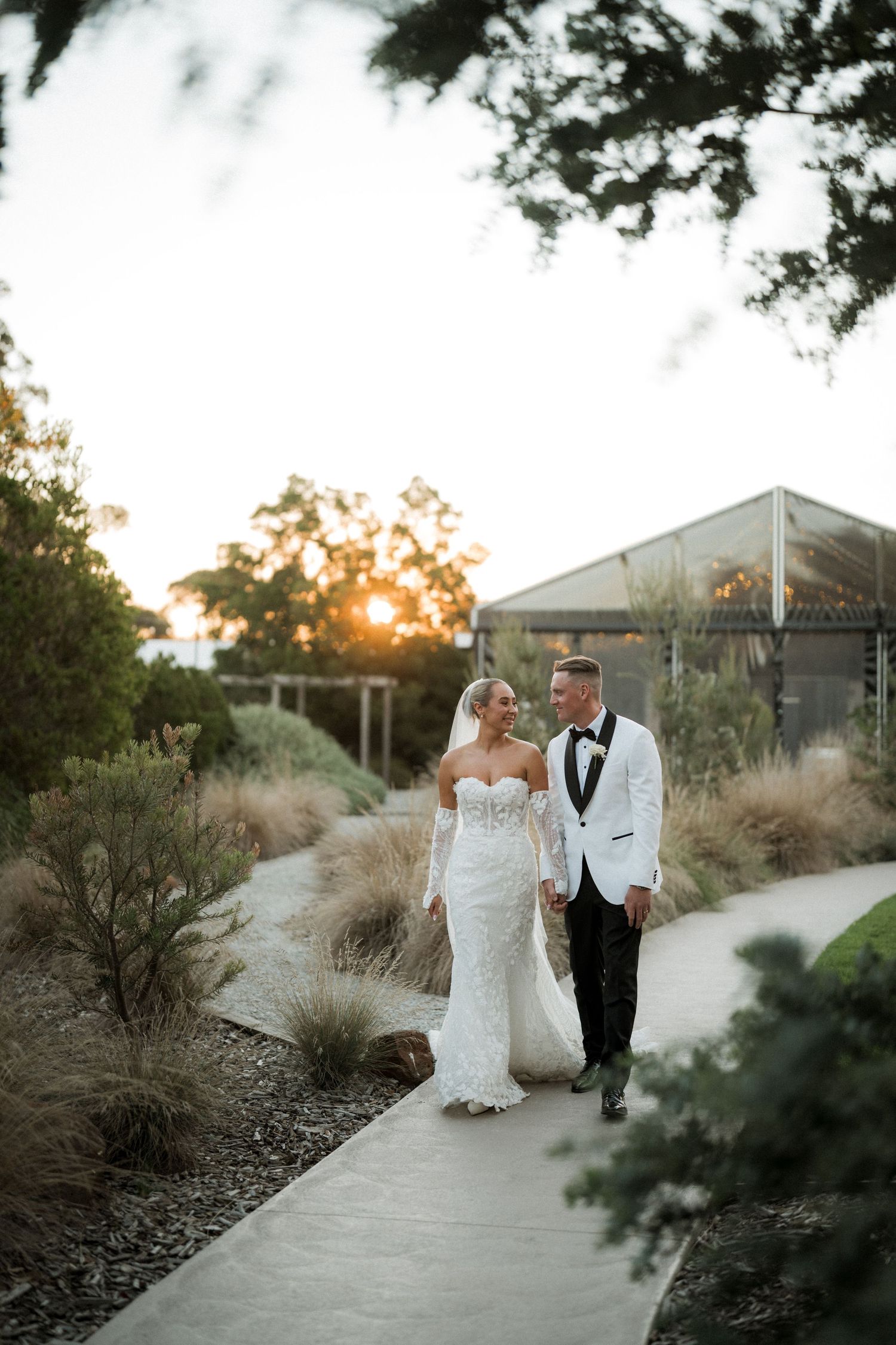 A couple walks along a garden path at sunset, with the bride in lace and groom in white tuxedo jacket.