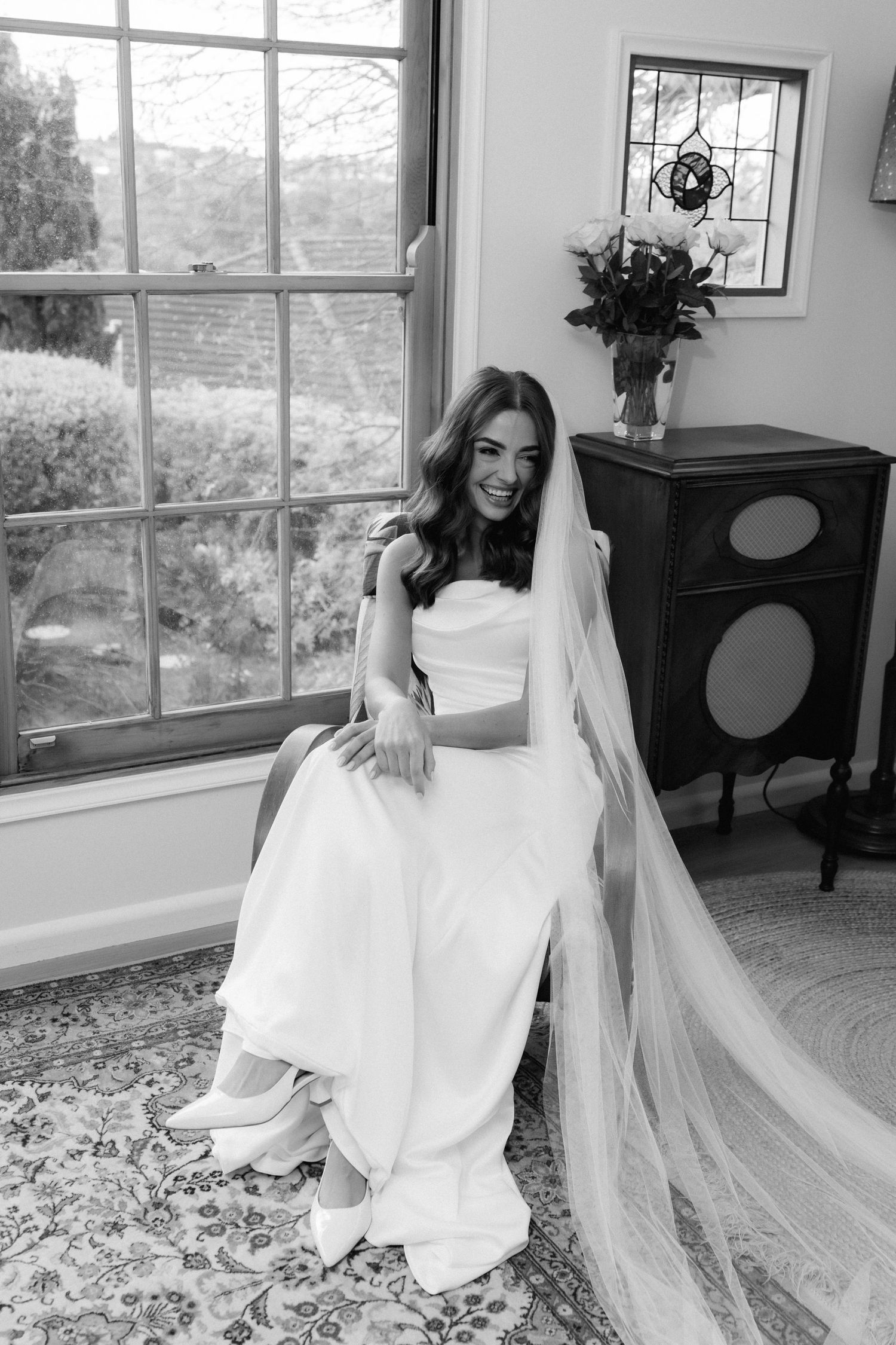 Black and white wedding photo showing a bride in an elegant white gown and veil seated by a large window.