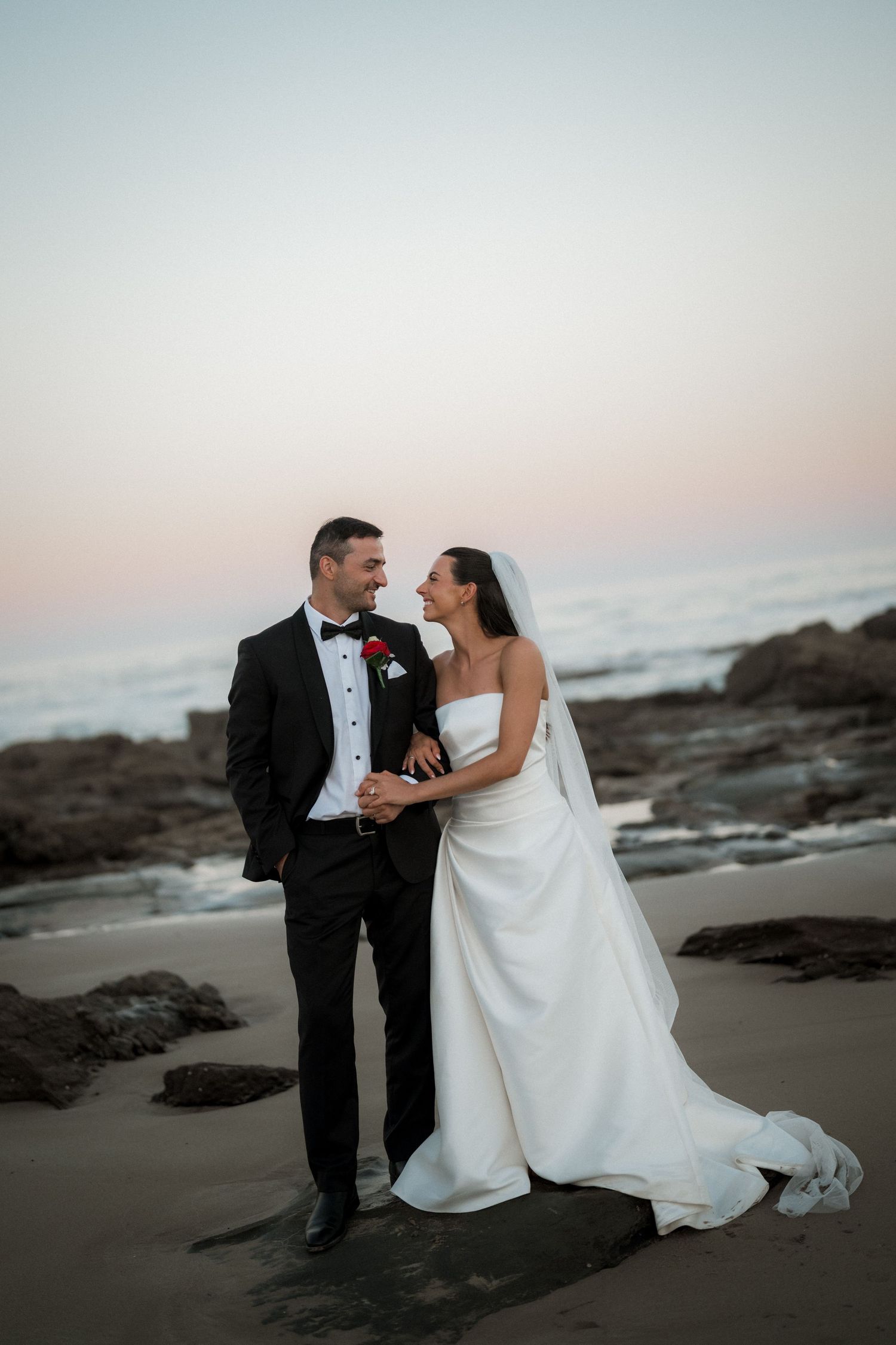Couple in wedding attire share intimate moment on rocky beach at sunset.