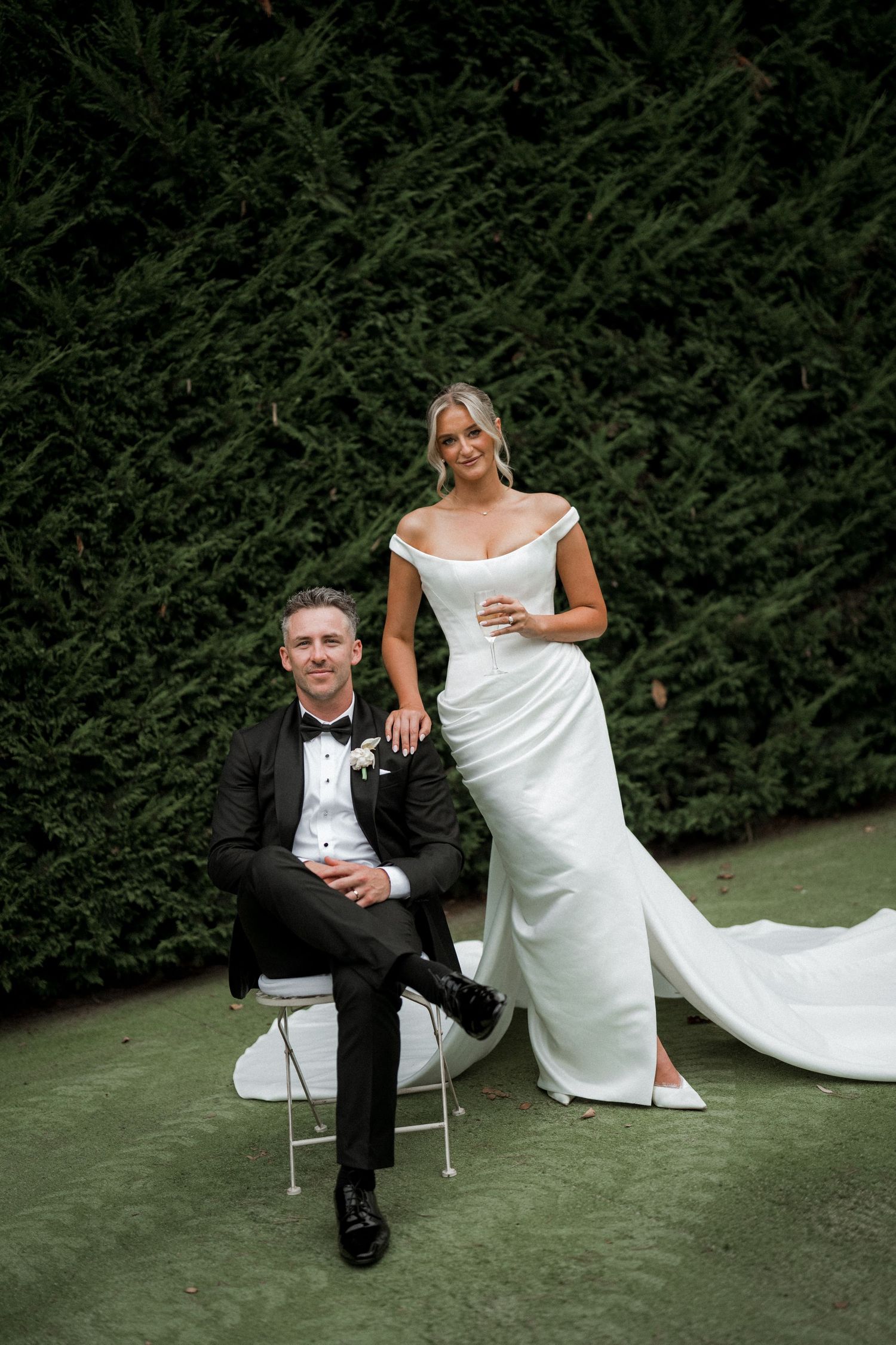 A couple in wedding attire poses casually on chairs outdoors with green hedges in the background.
