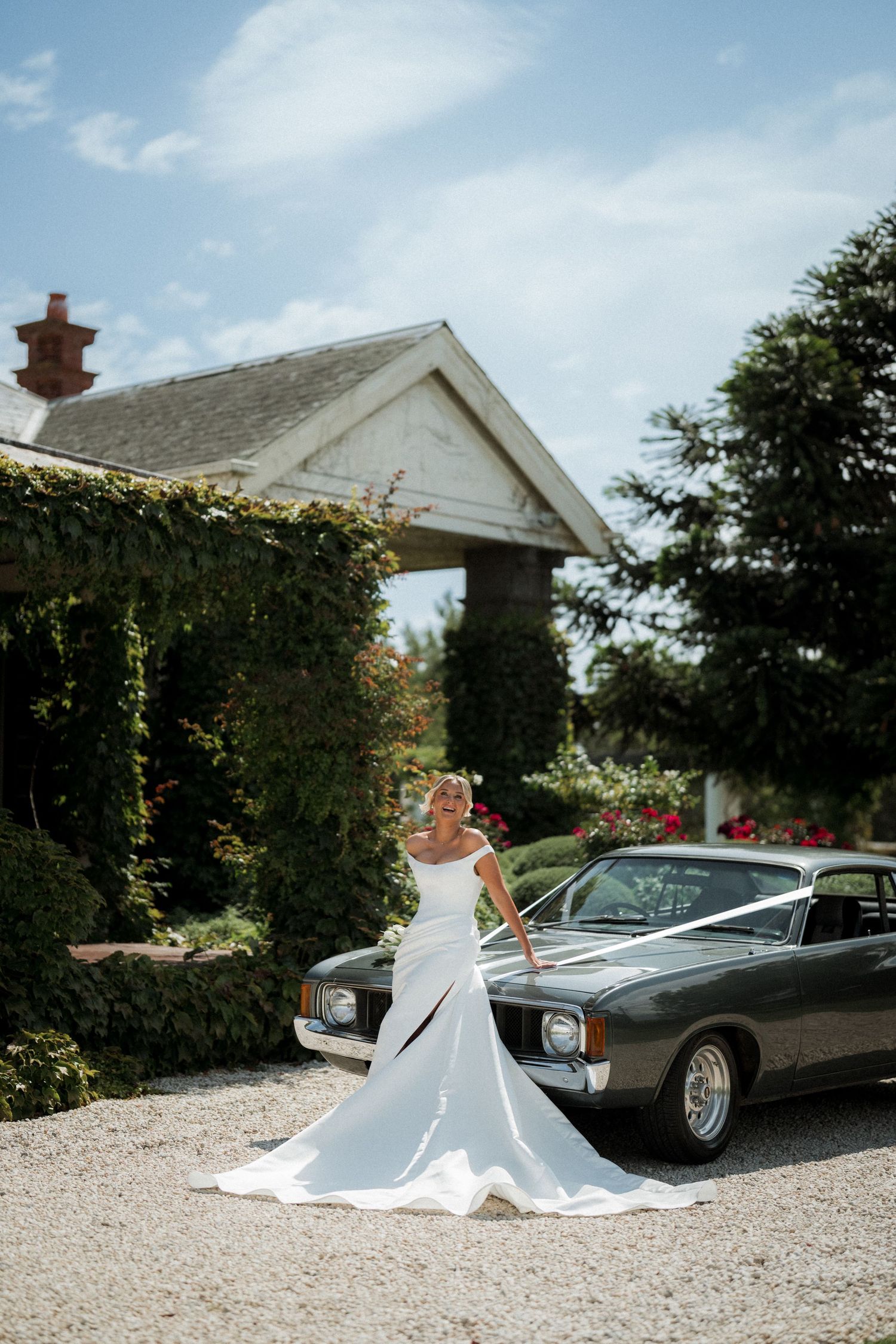 An elegant white wedding dress flowing on a gravel driveway with a classic car in the background.