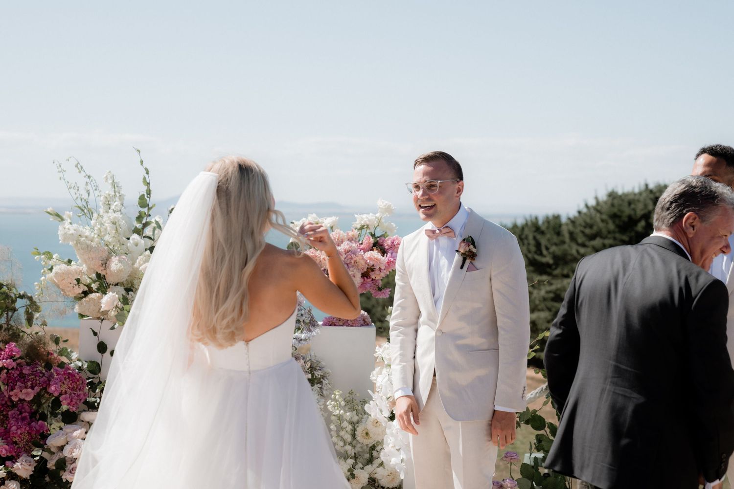 Wedding ceremony guests gather outdoors on a sunny day with ocean views and floral decorations in the background.