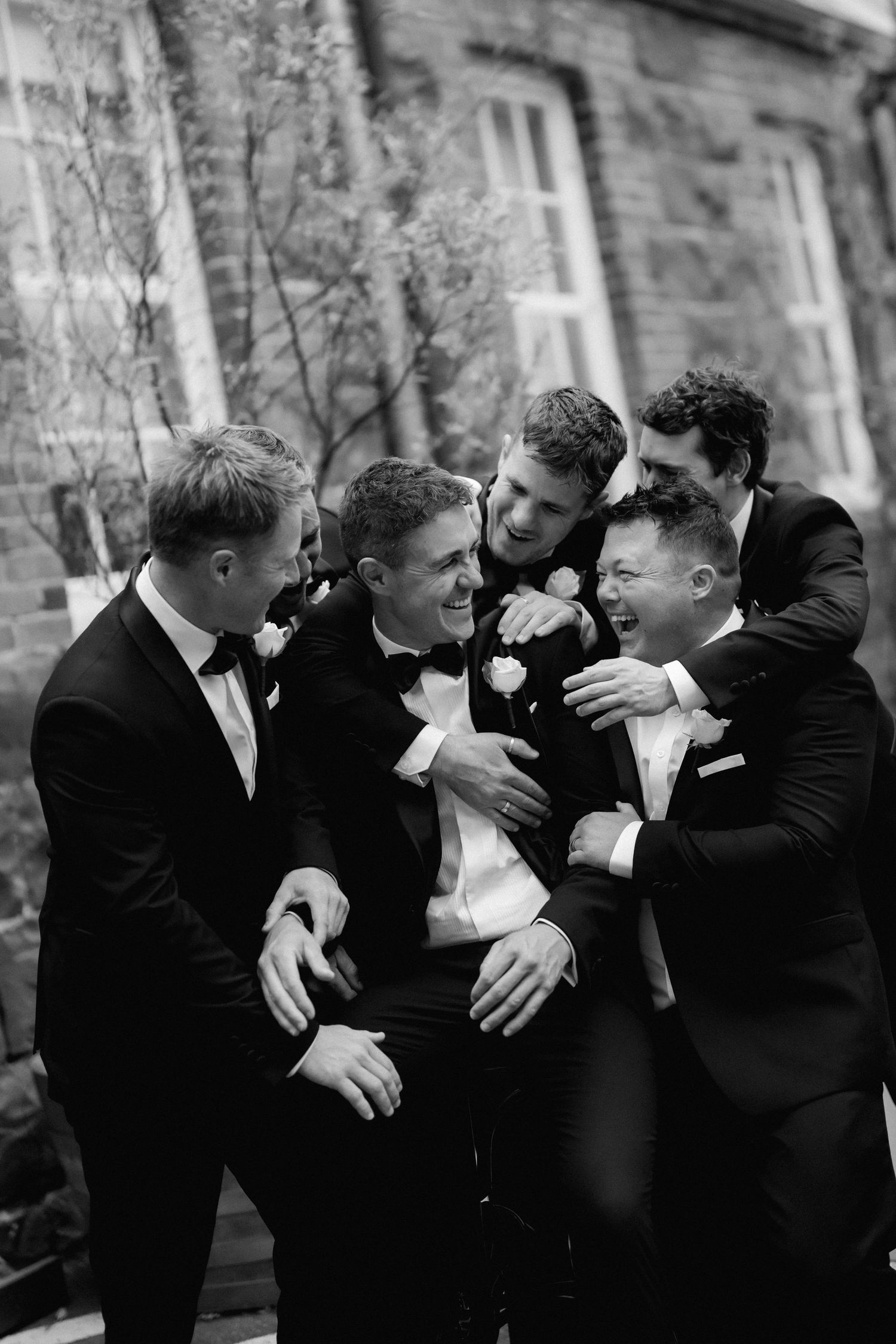 A candid black and white photo shows groomsmen in tuxedos laughing together during a wedding celebration.