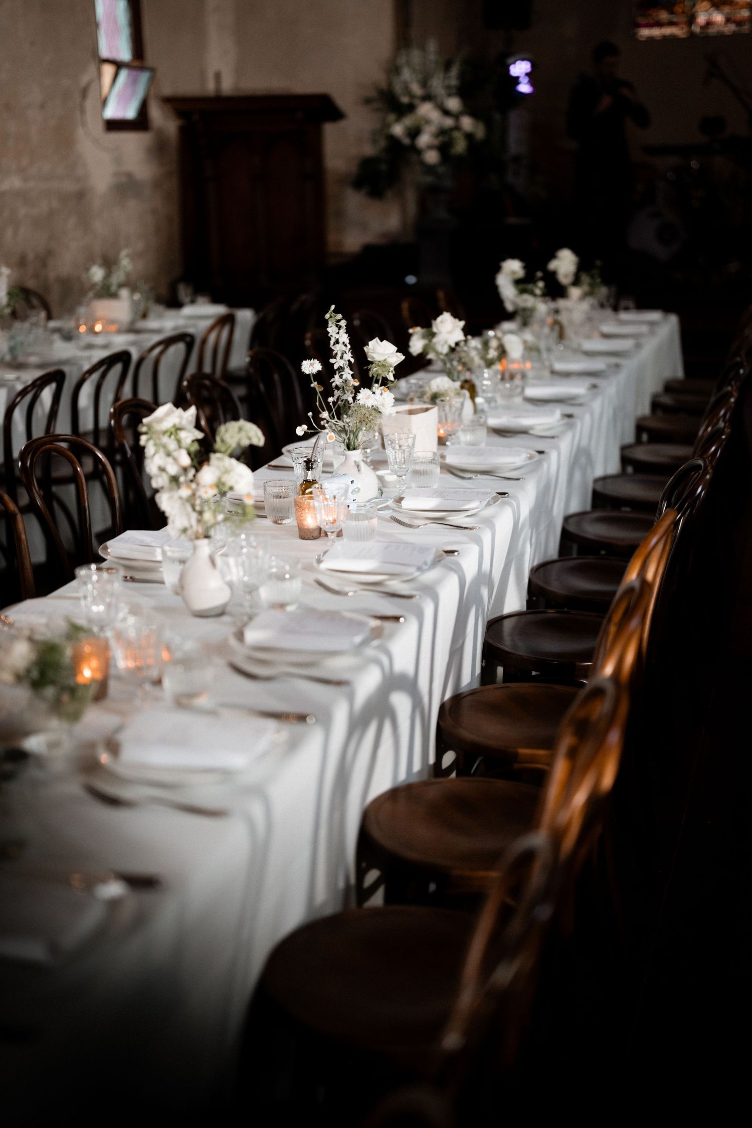 Elegant wedding reception table with white linens, dark wooden chairs, and delicate floral centerpieces in soft candlelight.