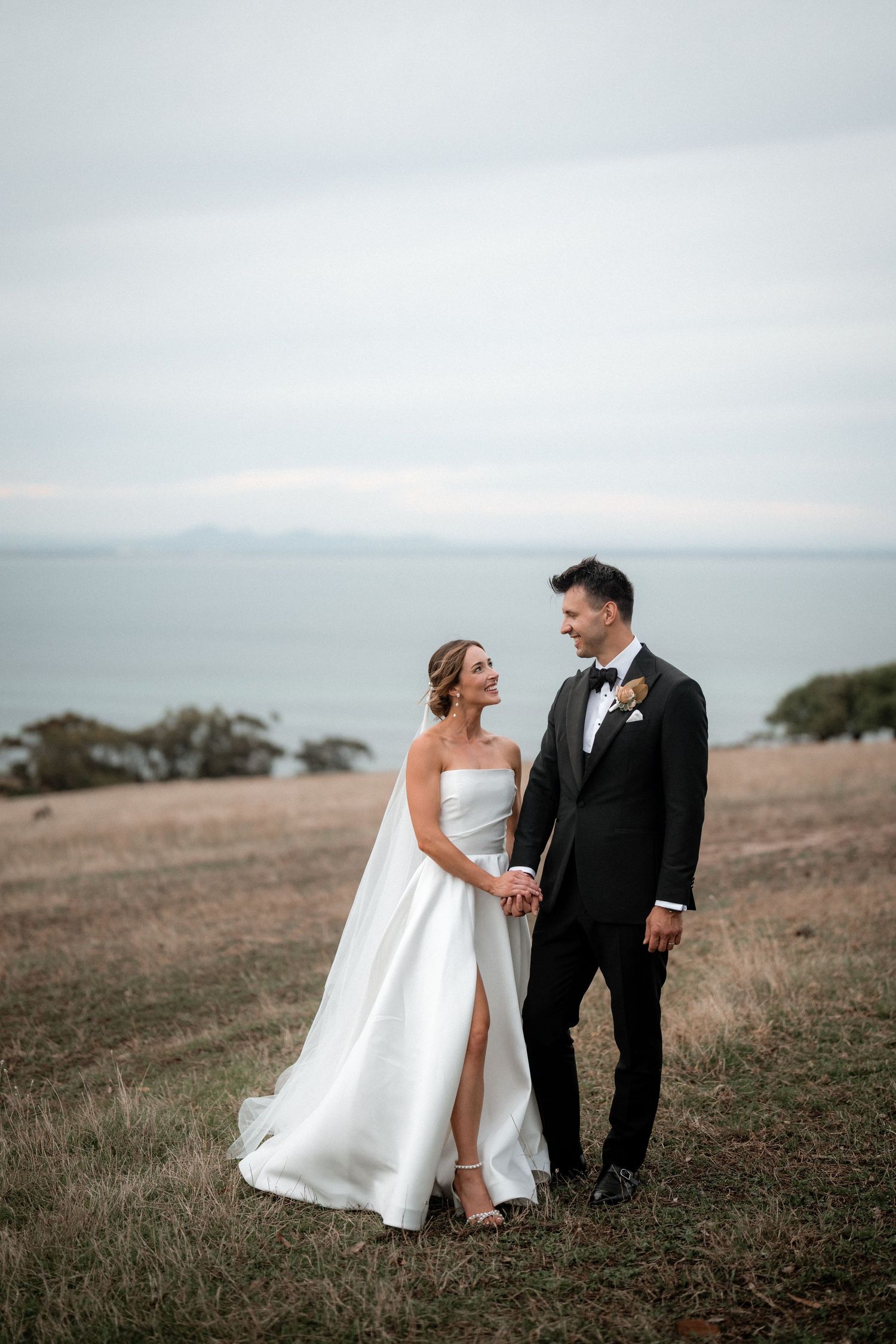 A couple in wedding attire stands on a hilltop overlooking a foggy ocean in the distance.
