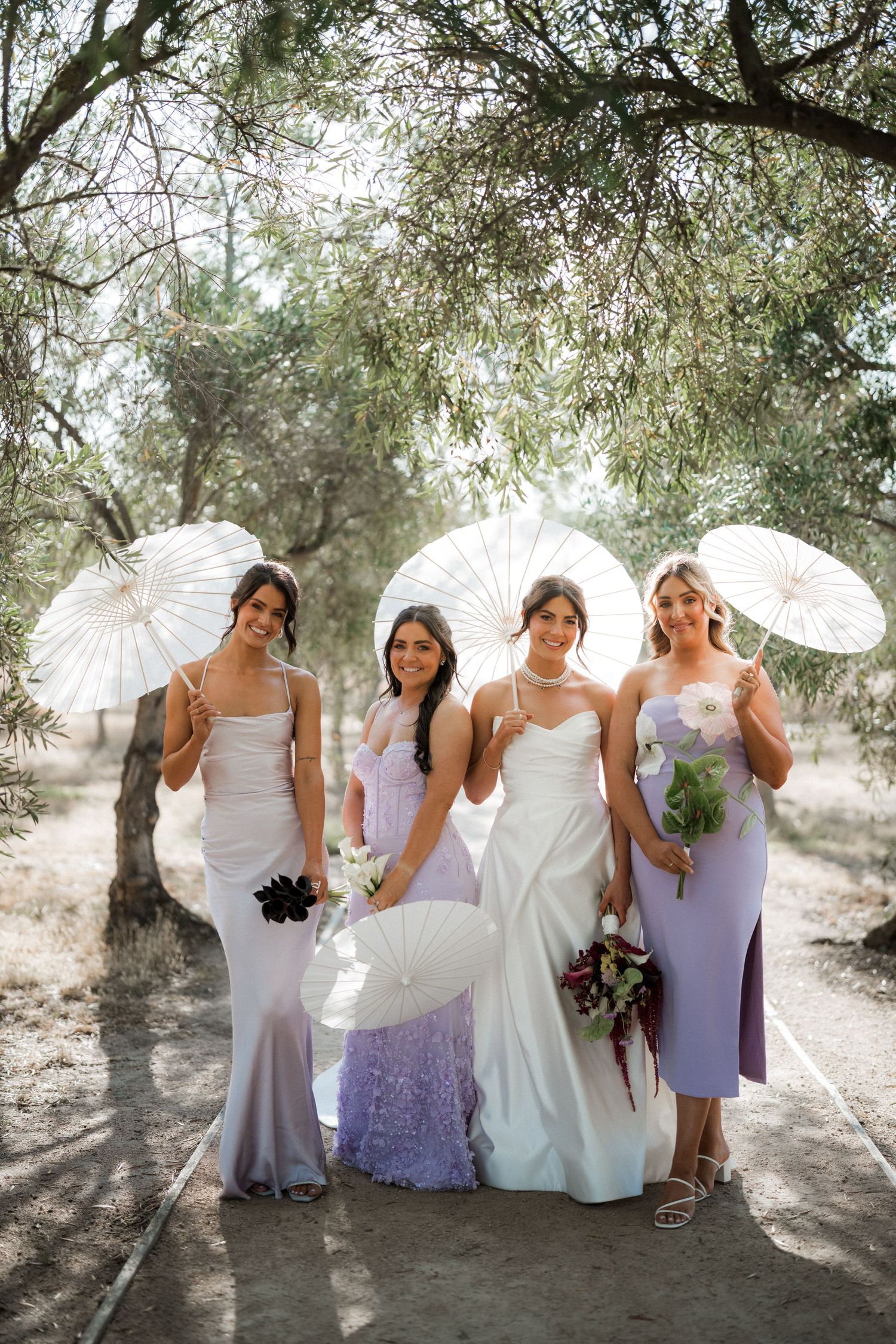 Bridal party in purple and white dresses posing with parasols in natural sunlight.
