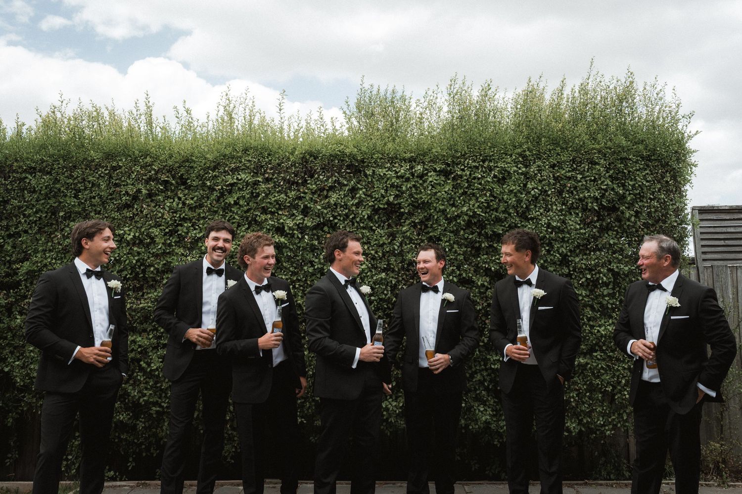 Groomsmen in black tuxedos pose together against a manicured hedge wall outdoors.