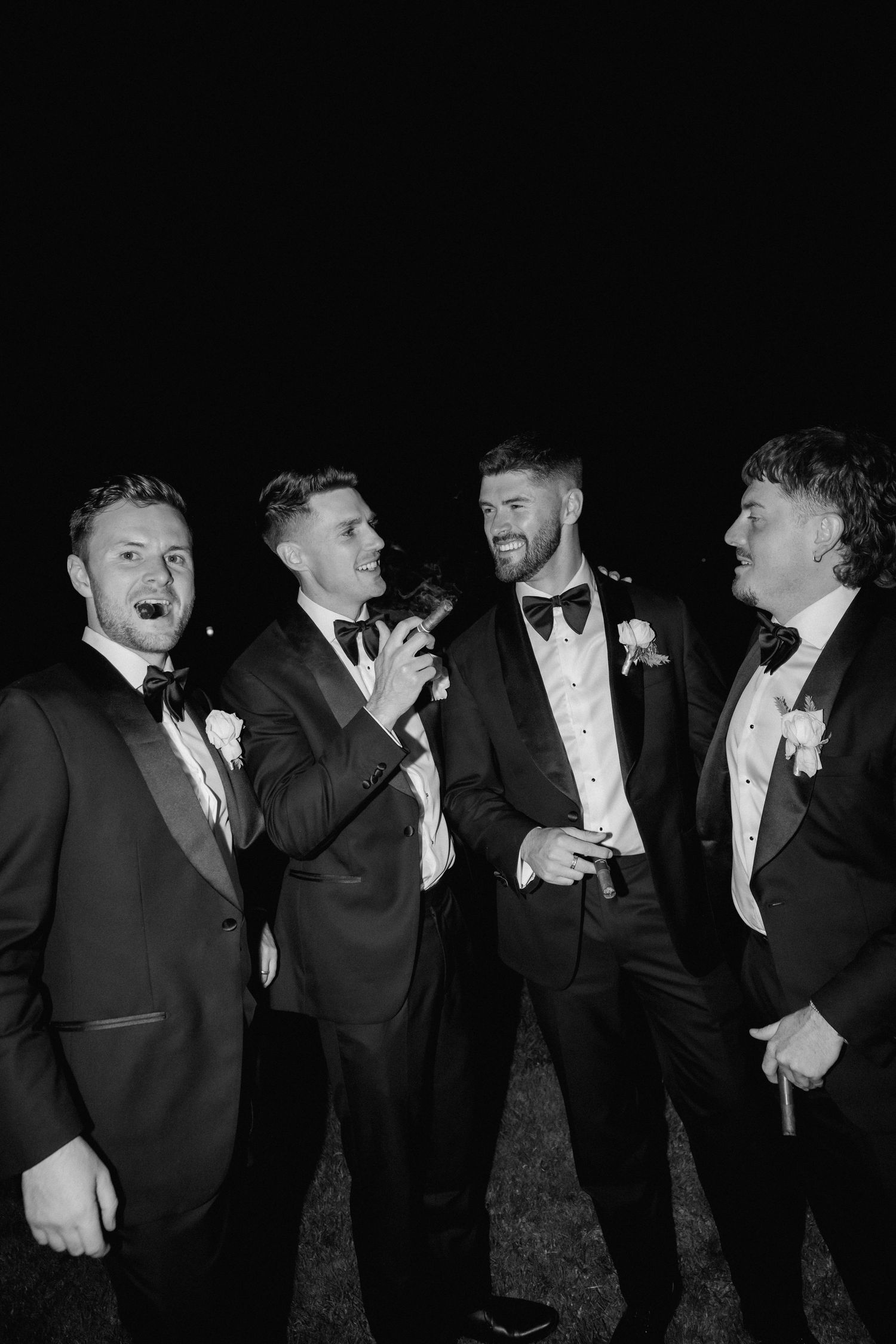 Groomsmen in tuxedos laughing and joking together in a candid black and white photo.