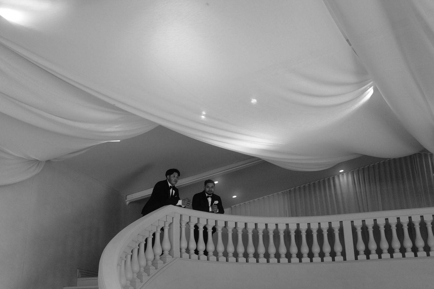 Two men in dark suits stand on an ornate balcony with a white balustrade beneath draped fabric ceiling swags in a black and white photo.