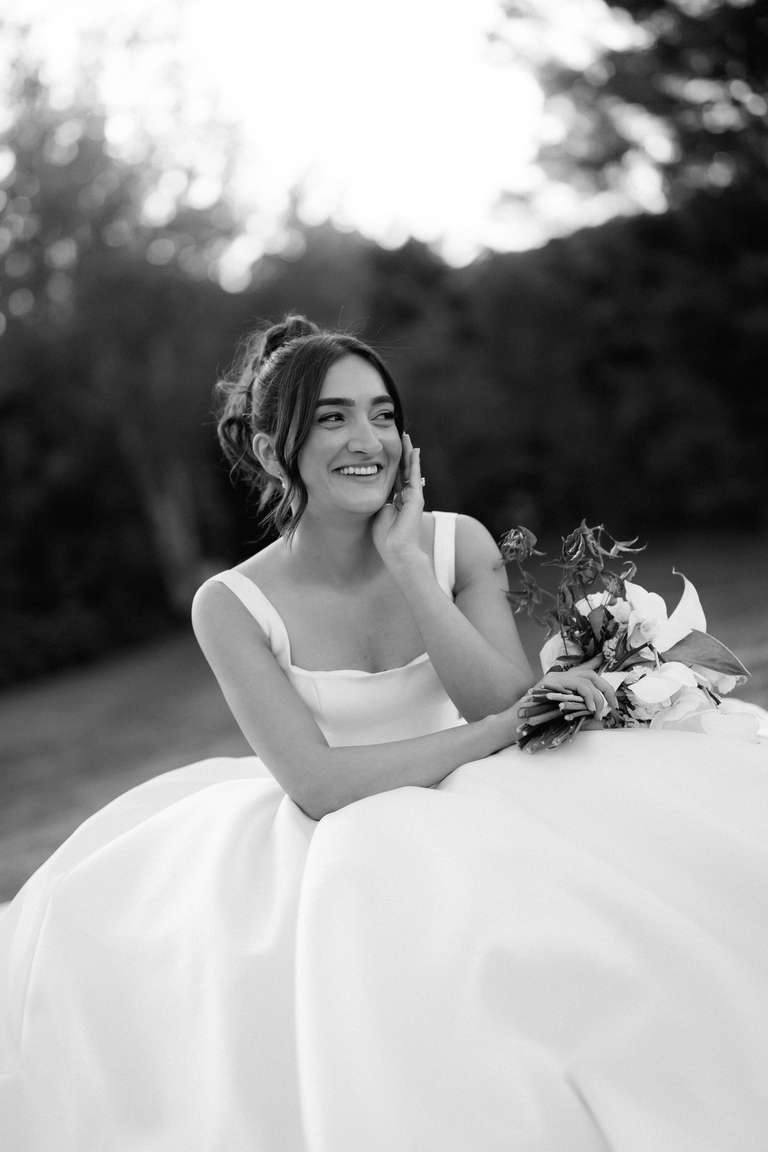 Black and white portrait of a joyful bride in a square-neck wedding gown holding a bouquet and smiling outdoors.