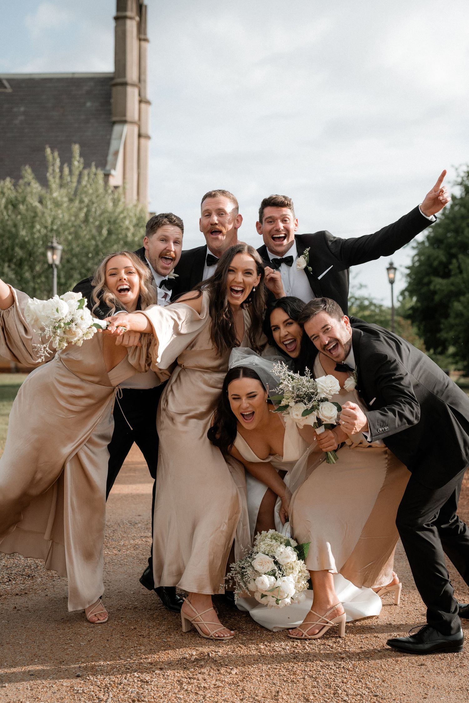Wedding party poses for fun group photo in champagne and black attire with white floral bouquets.