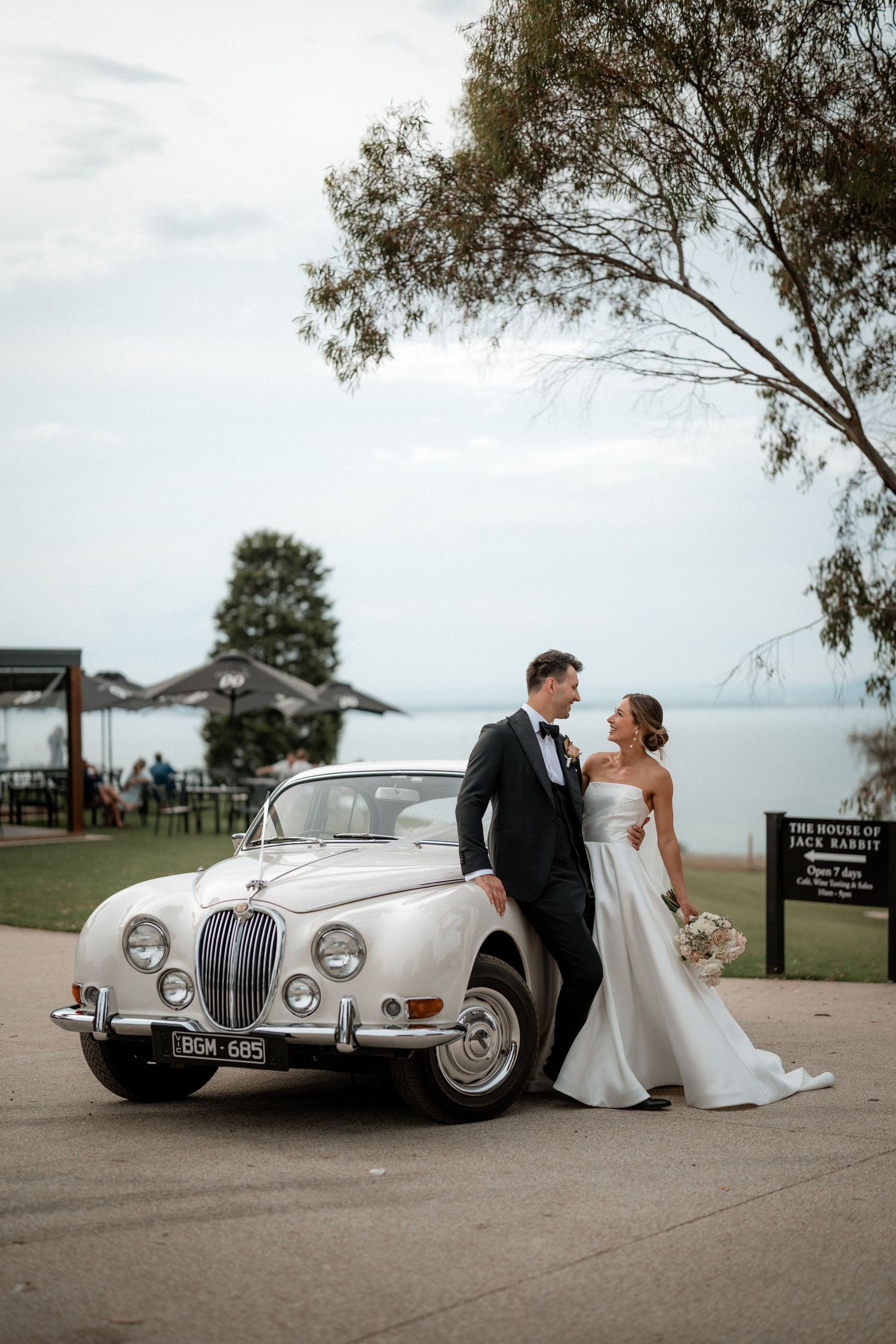 Classic silver vintage car parked on paved ground with trees and cloudy sky at an outdoor wedding venue.