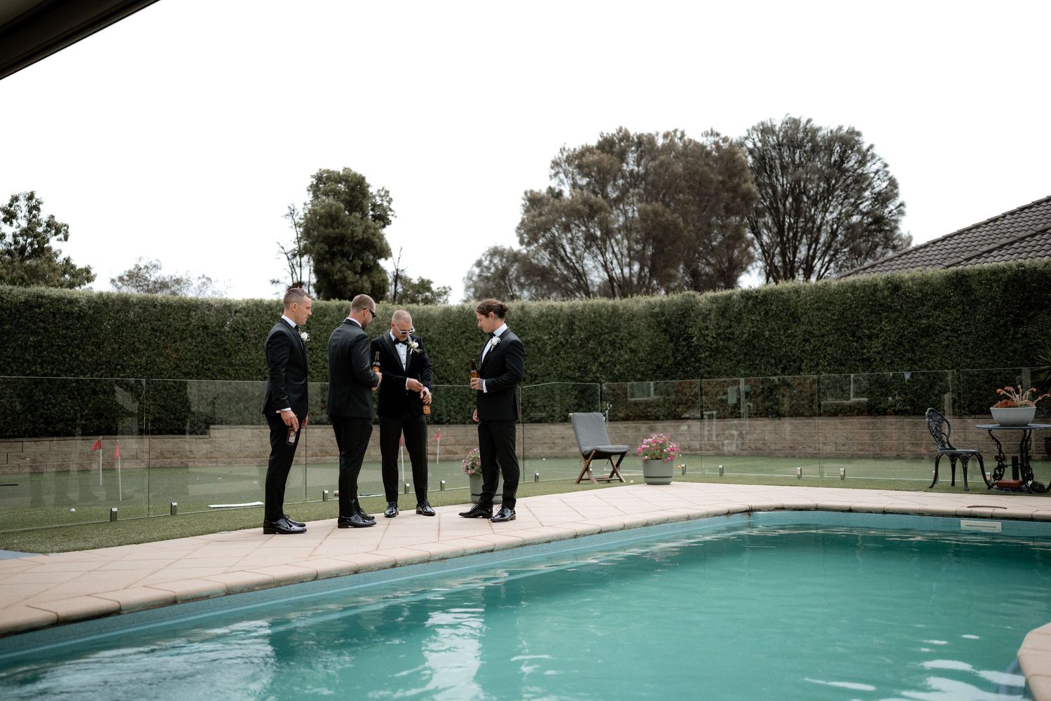 A group of formally dressed men stand beside a swimming pool surrounded by hedges during a wedding event.