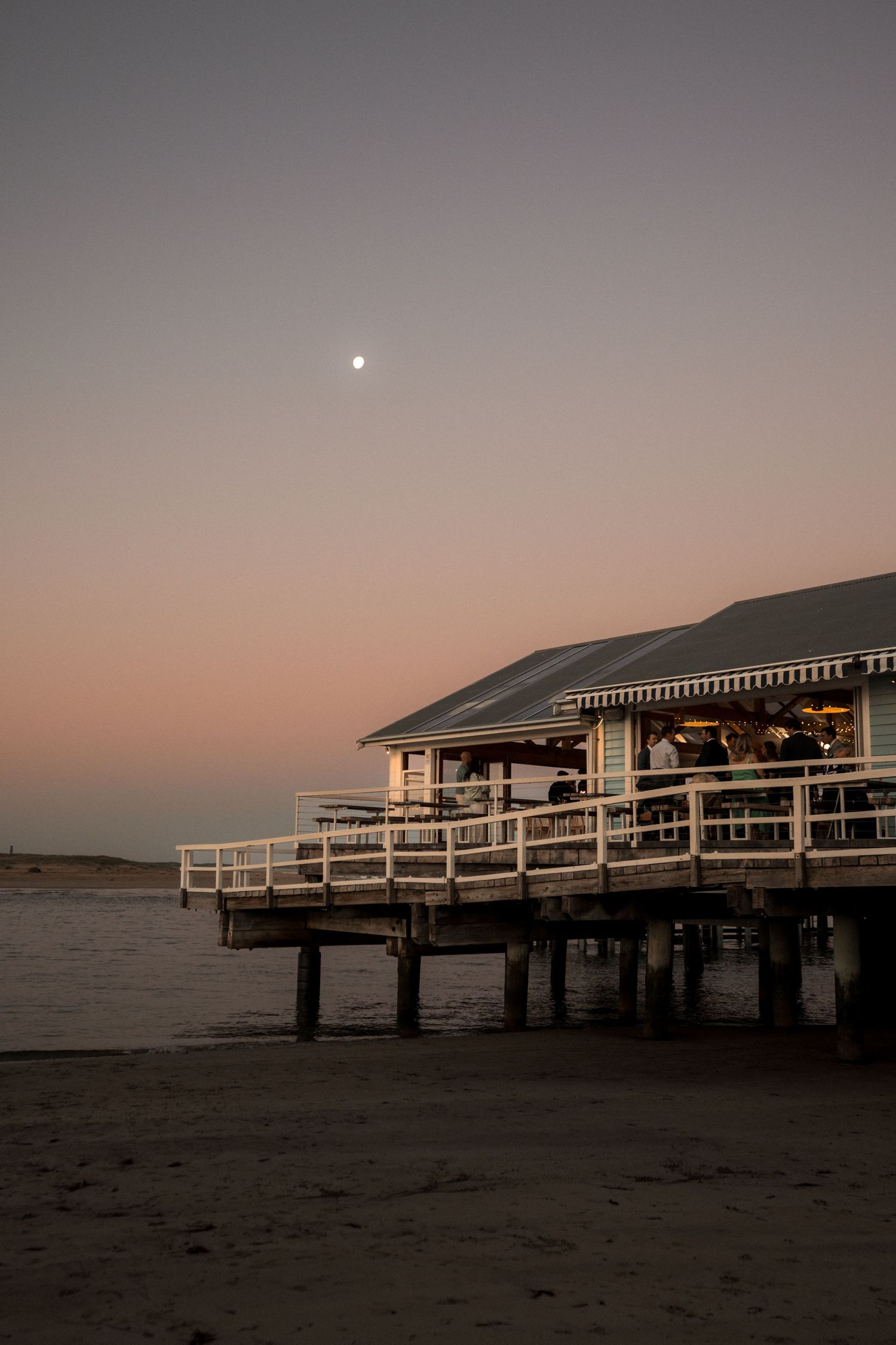 A wooden pier restaurant glows at dusk with a full moon rising over the sandy beach and ocean.