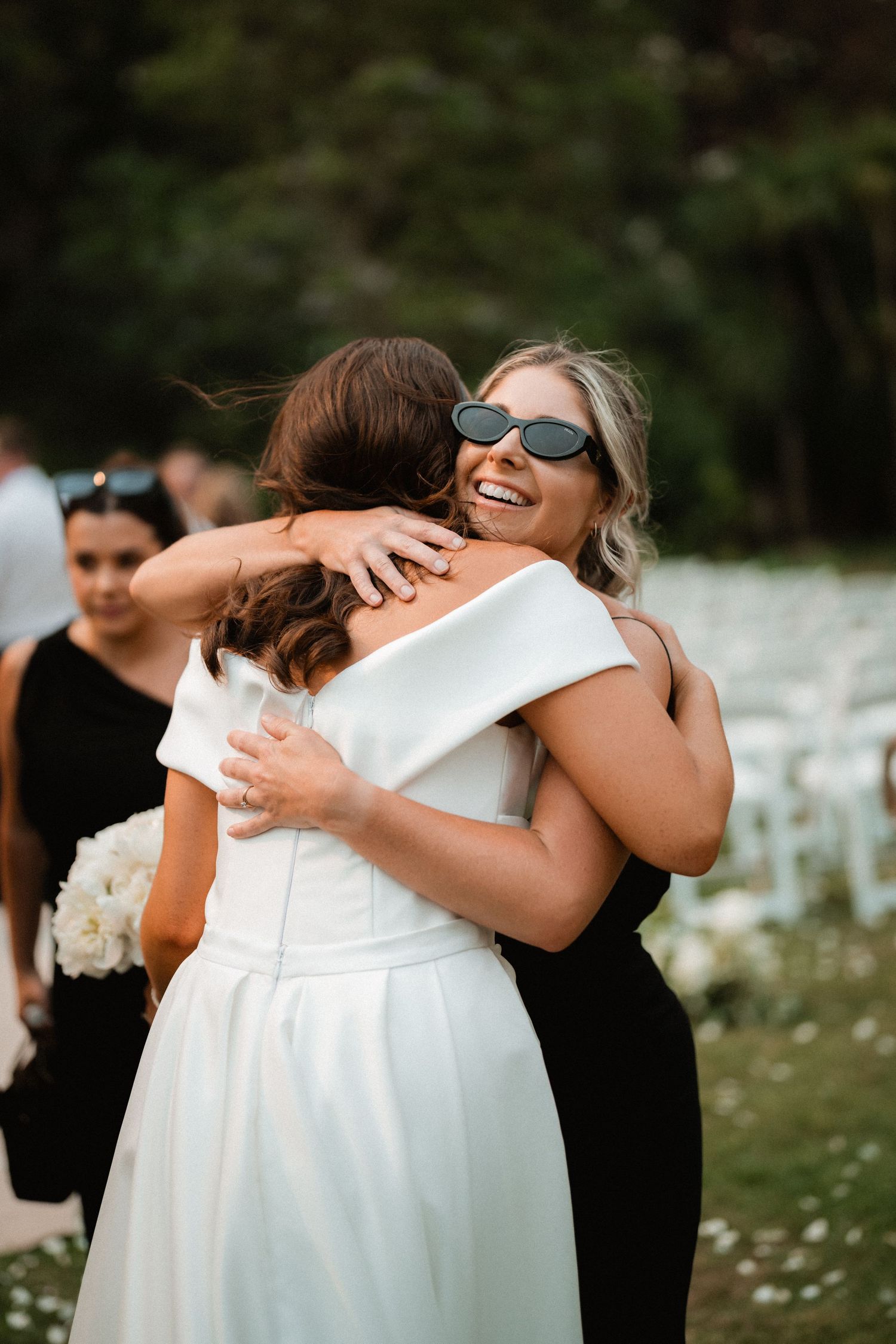 Two wedding guests share an emotional embrace outdoors with white chairs and flowers in the background.