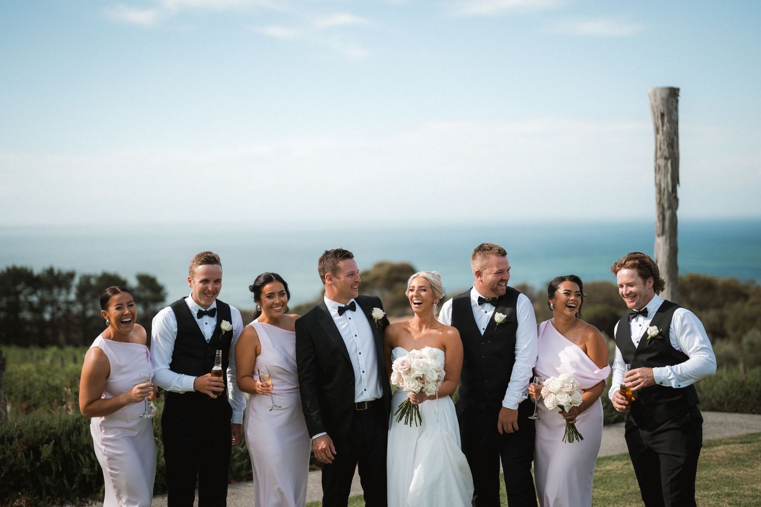 A wedding party poses together against an ocean backdrop with blue skies.