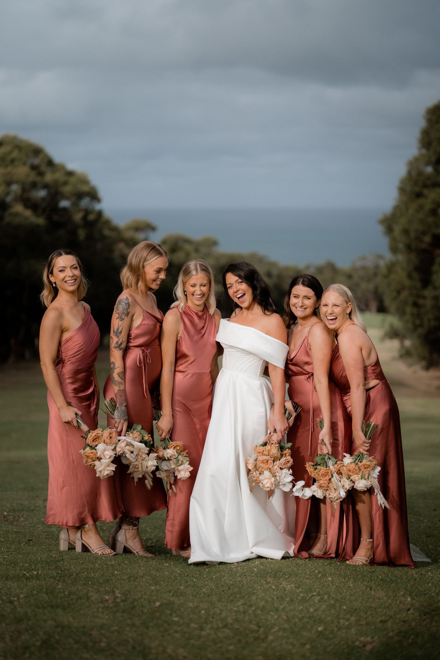 A bridal party in coral pink bridesmaid dresses and white wedding gown pose together on a grassy hillside.