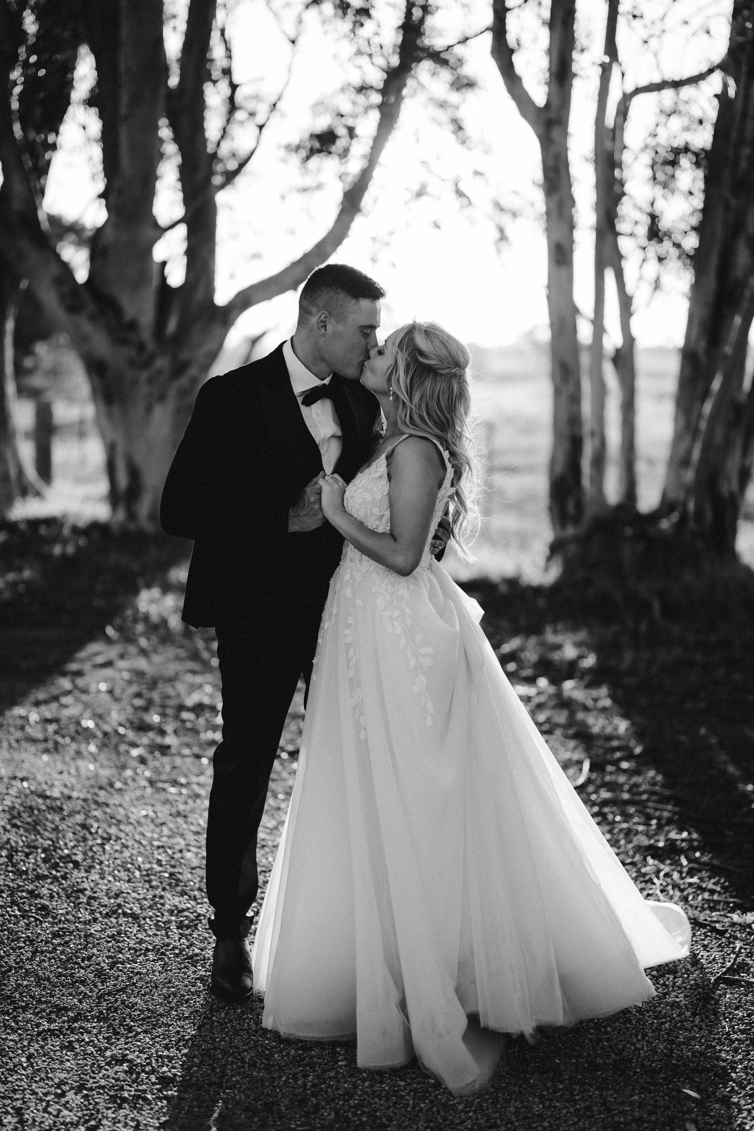 Black and white romantic wedding photo showing a couple in formal attire embracing on a tree-lined path.