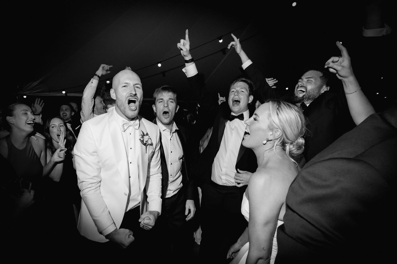 A black and white photo of wedding guests dancing and celebrating with enthusiastic expressions at a reception party.