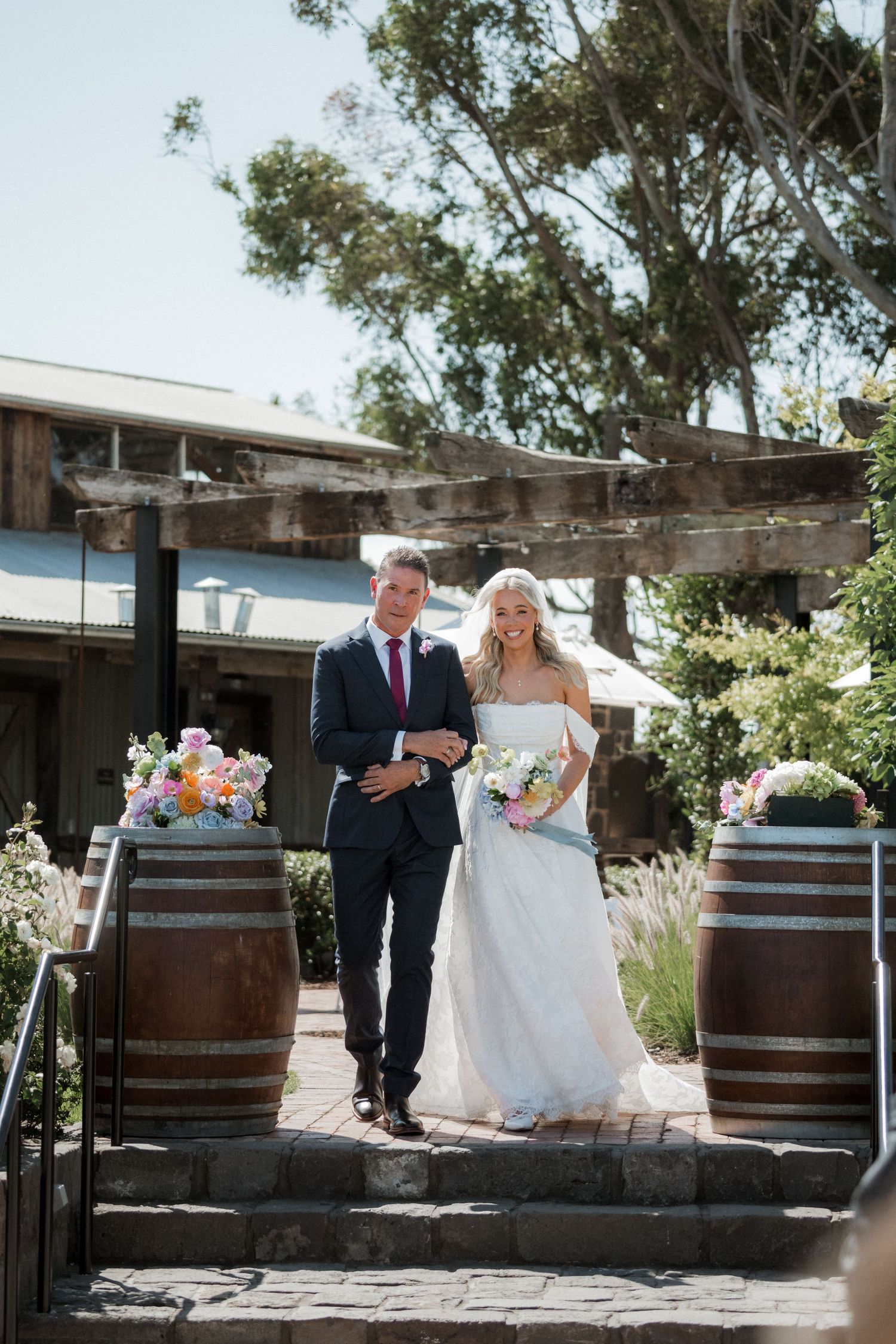Bride in white off-shoulder gown walks with escort past wine barrels at an outdoor winery wedding venue.