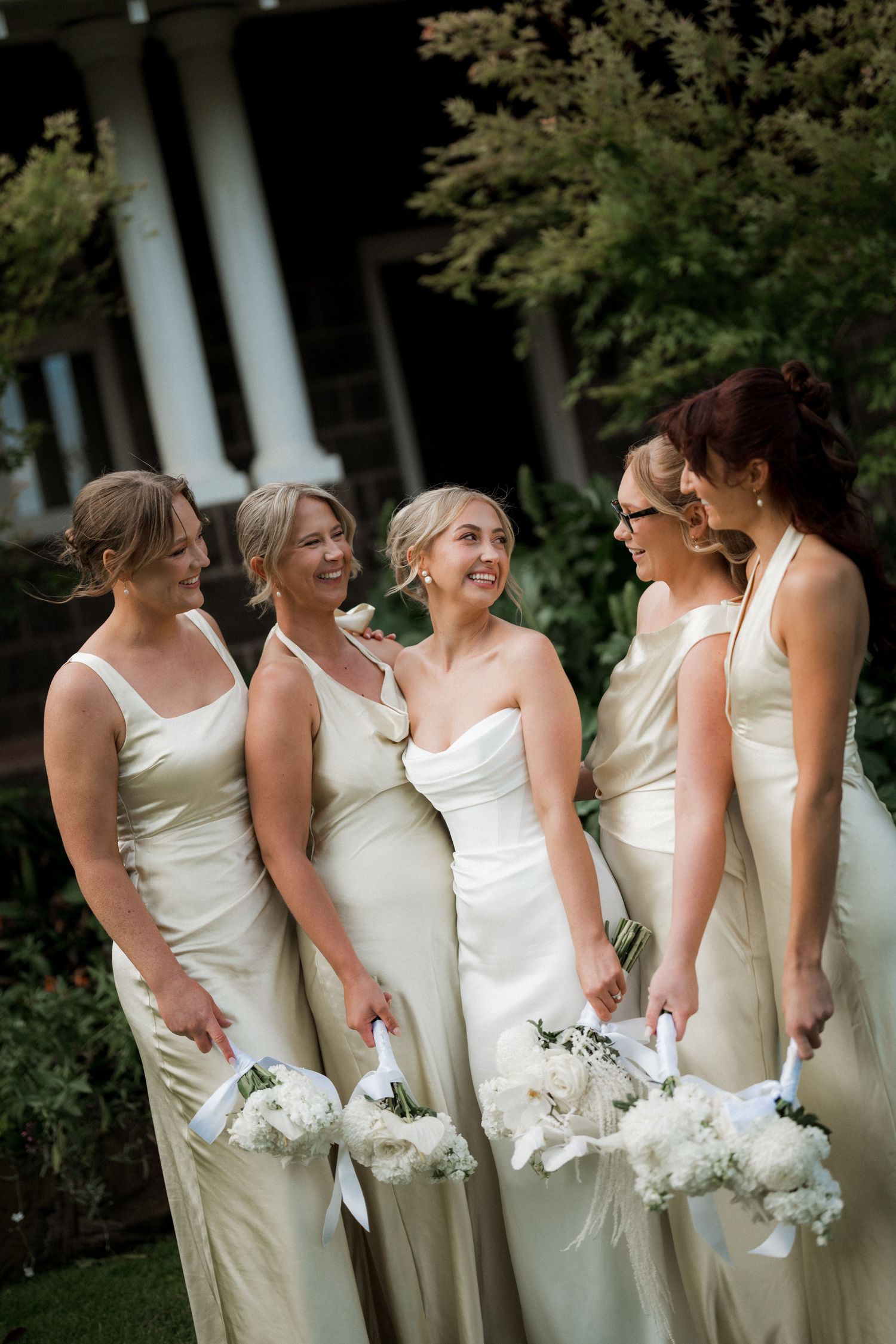 Bridesmaids in cream-colored dresses hold white bouquets while standing in a row.