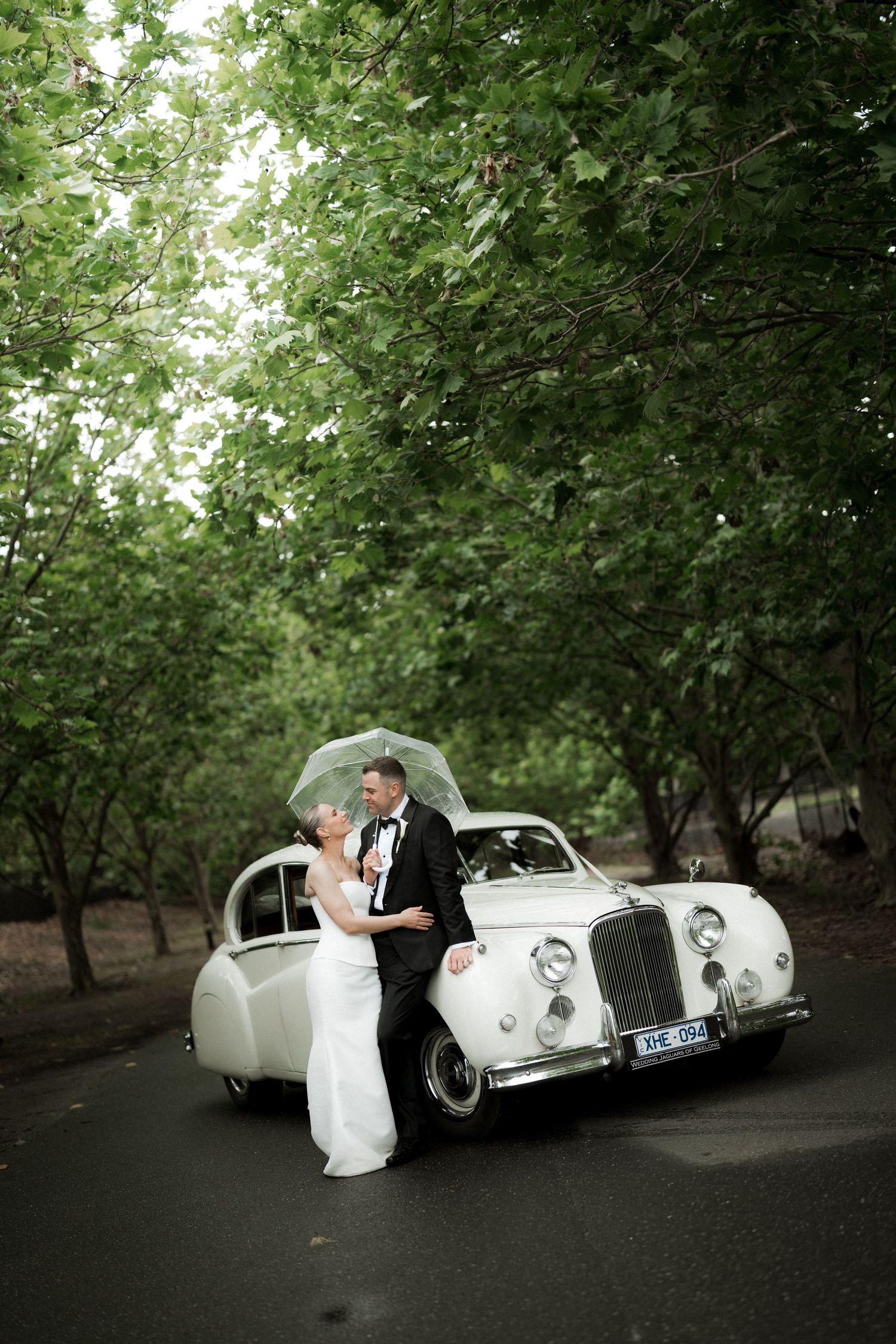 Classic white vintage car provides elegant backdrop for wedding photos in tree-lined setting.