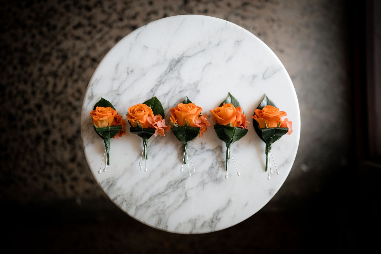 Five orange roses arranged in a row on a round white marble surface against a dark background.