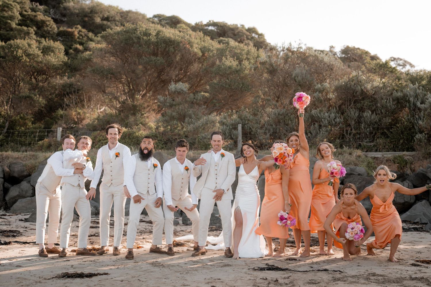 Wedding party arranged in line on beach wearing white and peach colored outfits.