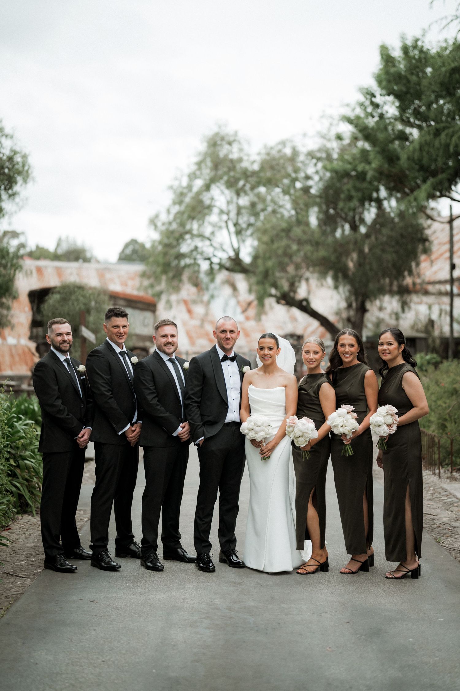 Wedding party in black and white attire poses together on a paved pathway with trees in the background.
