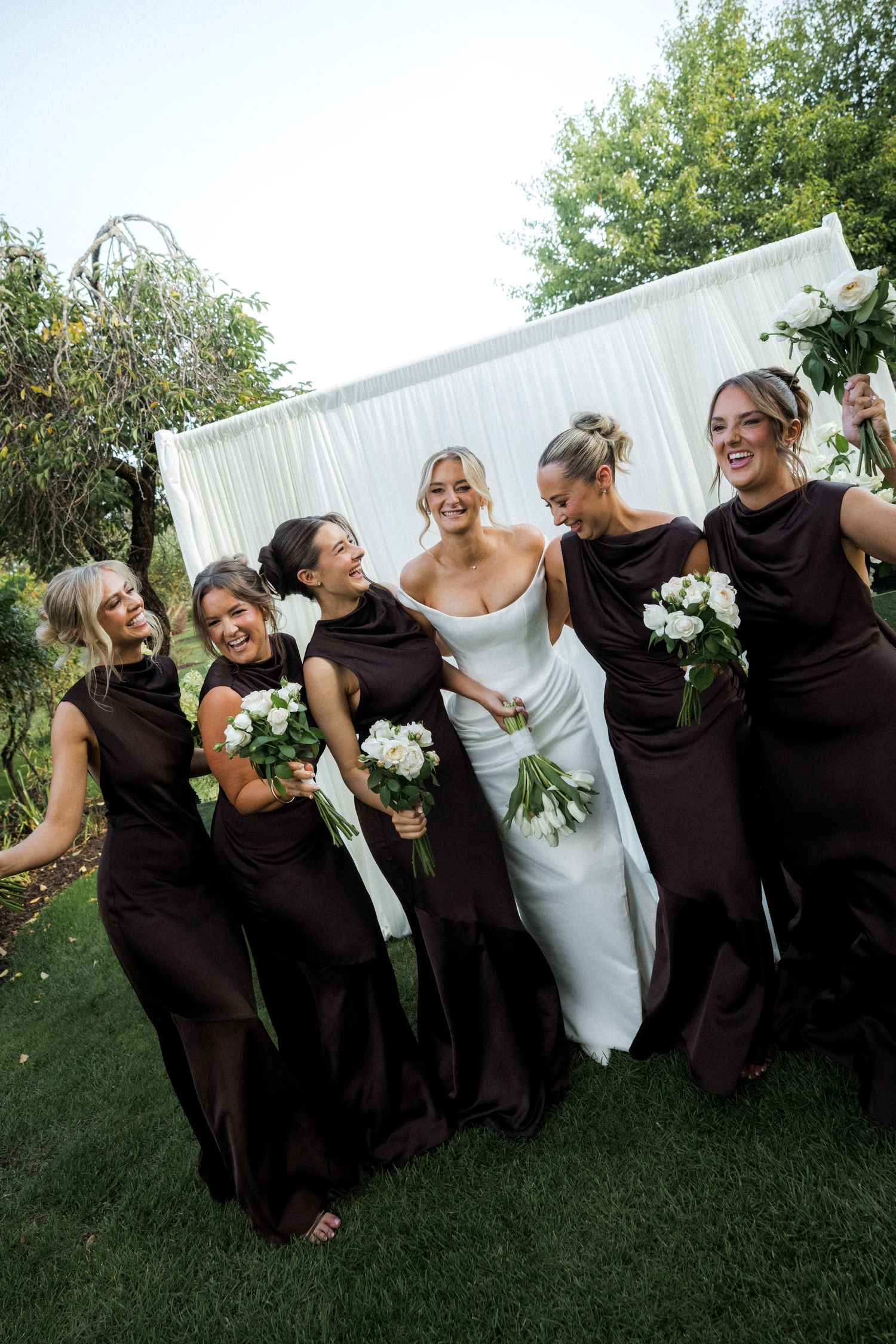 Wedding party strikes playful pose on grass with bridesmaids in black dresses.