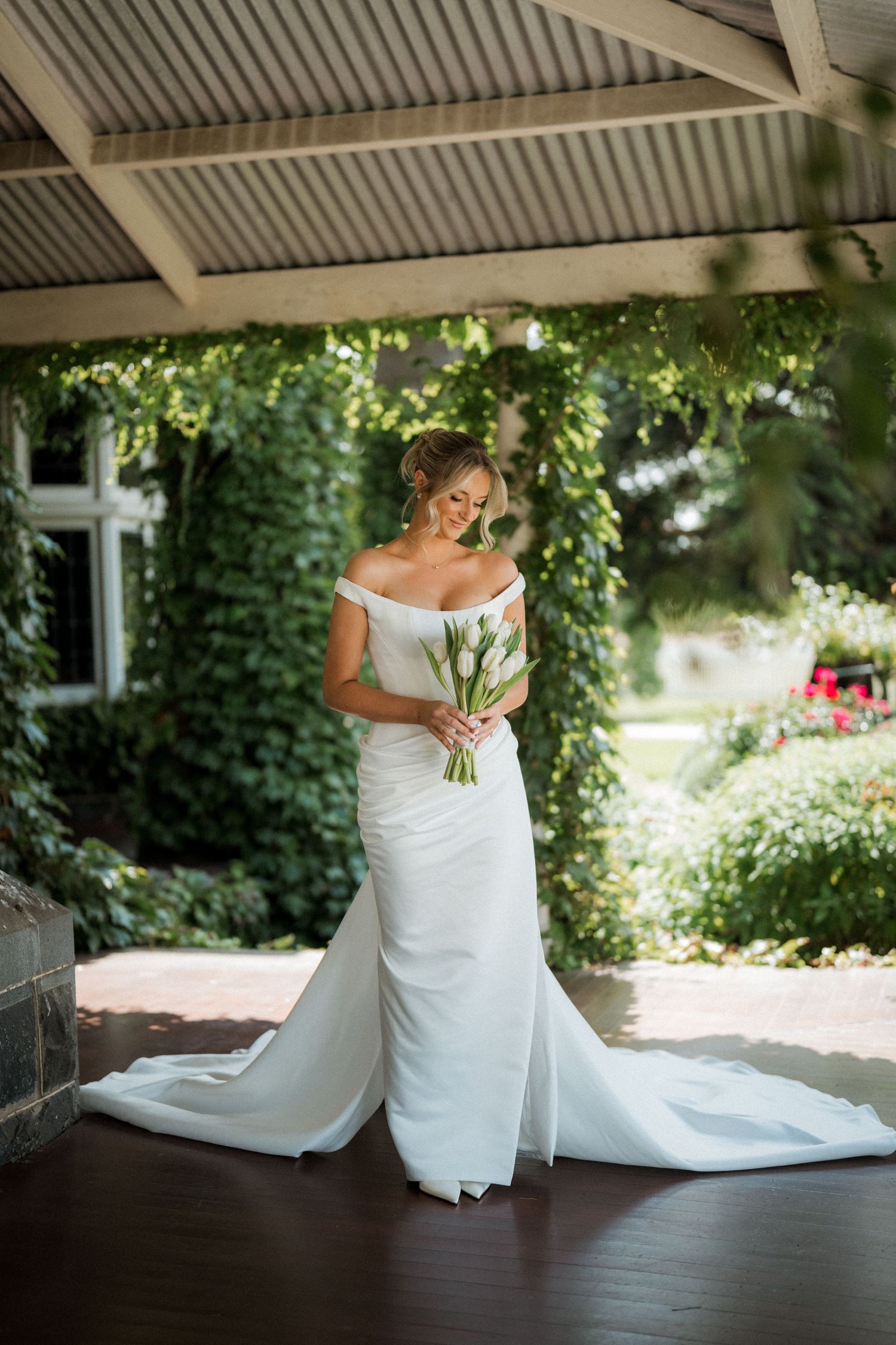 Bride in white gown sitting on wooden deck surrounded by greenery and natural light.