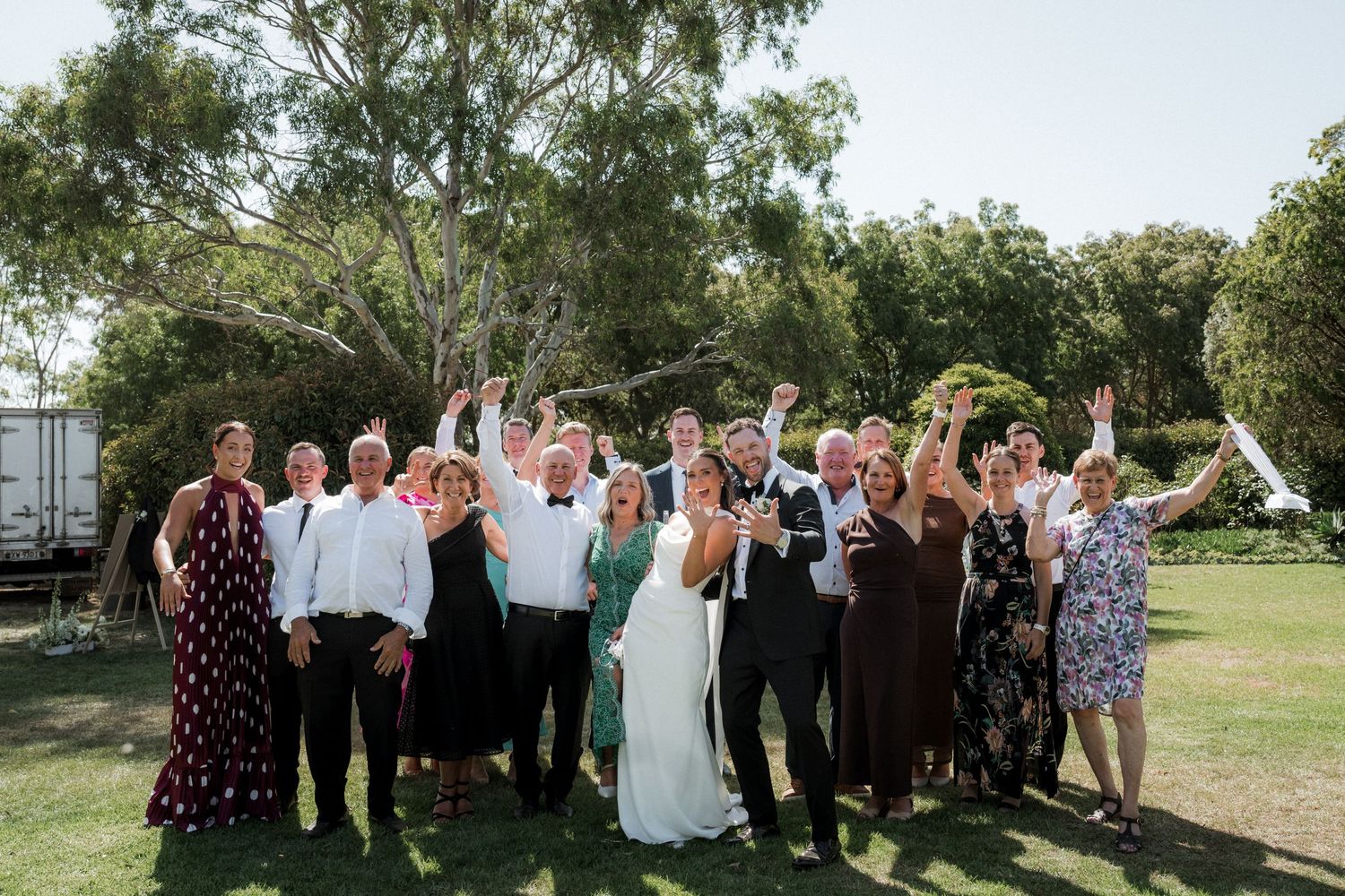 Wedding guests gather outdoors in a garden setting for a group photo on a sunny summer day.
