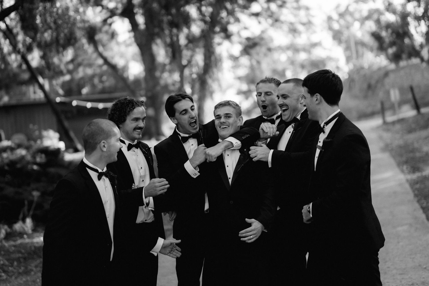 Groomsmen in black tuxedos laughing together outdoors during wedding photography session.