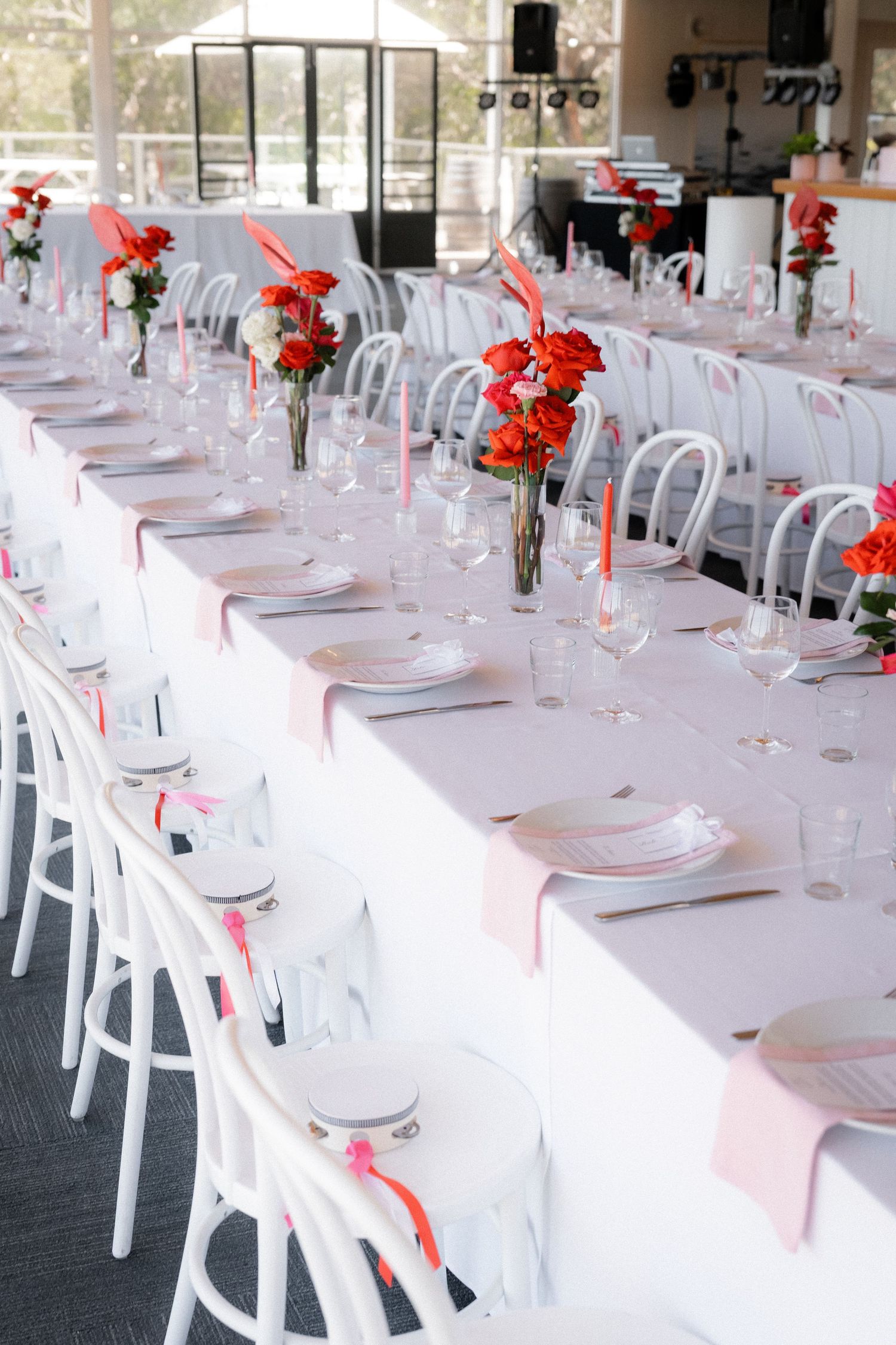 Elegant wedding reception with white chairs, long tables, pink linens, red floral centerpieces, and glassware in a bright venue.